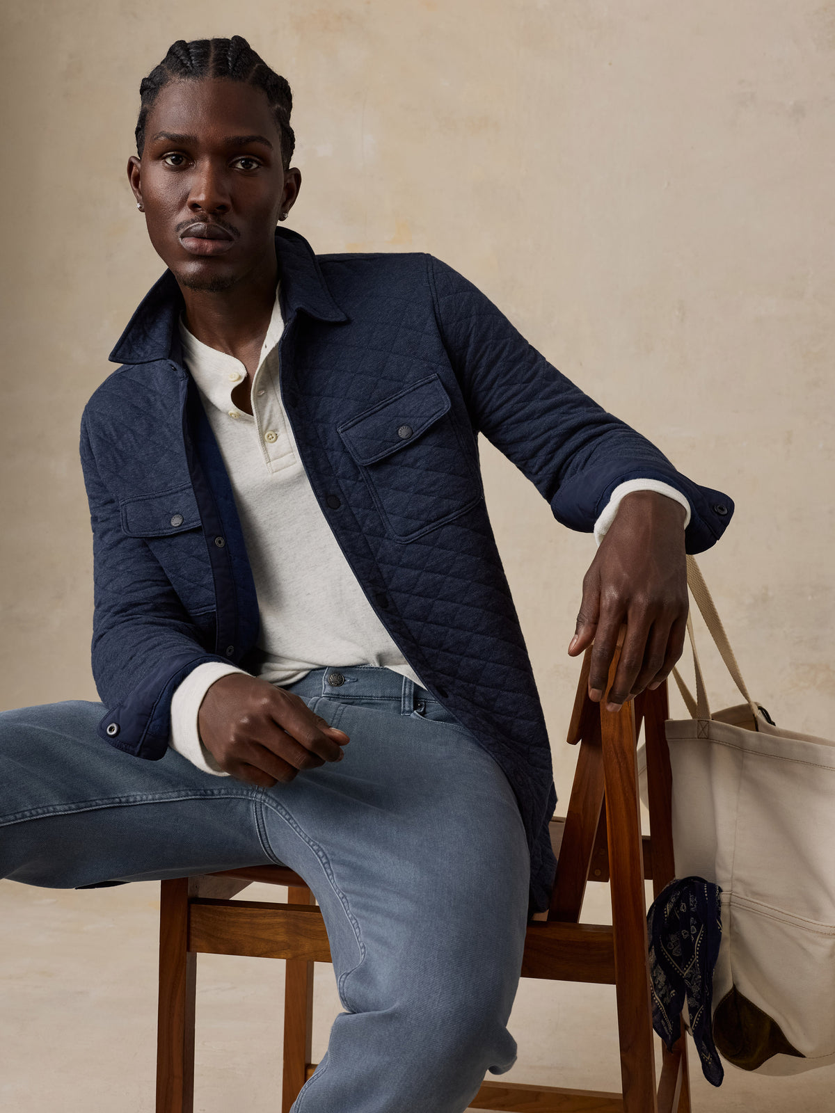 A man sits on a wooden chair wearing a navy quilted jacket and light blue jeans with a cream canvas tote bag resting on the chair against a warm beige studio backdrop., image 2 of 7