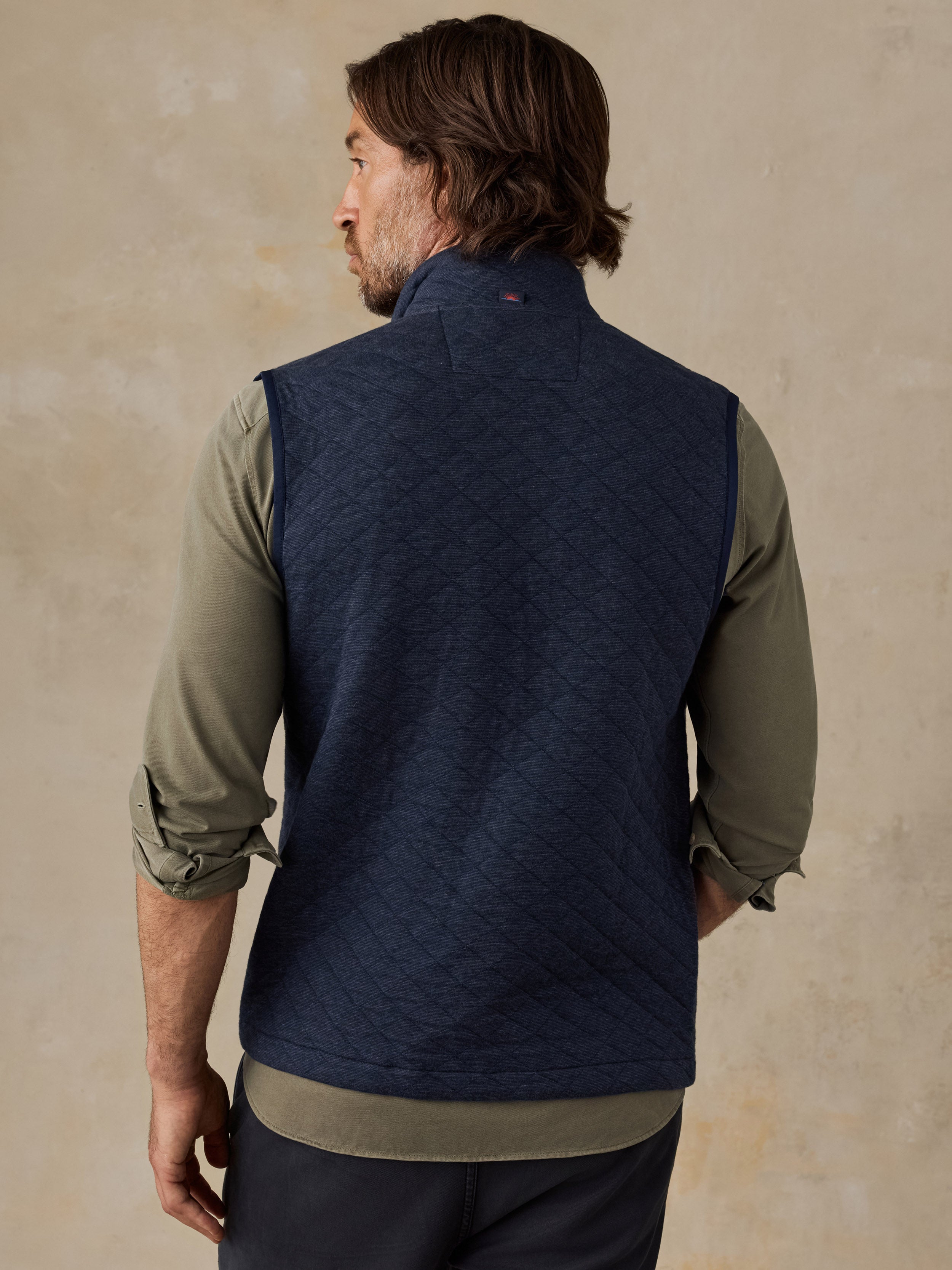 A man shown from the back wearing a navy blue quilted vest over an olive green long-sleeve shirt, set against a textured beige background.