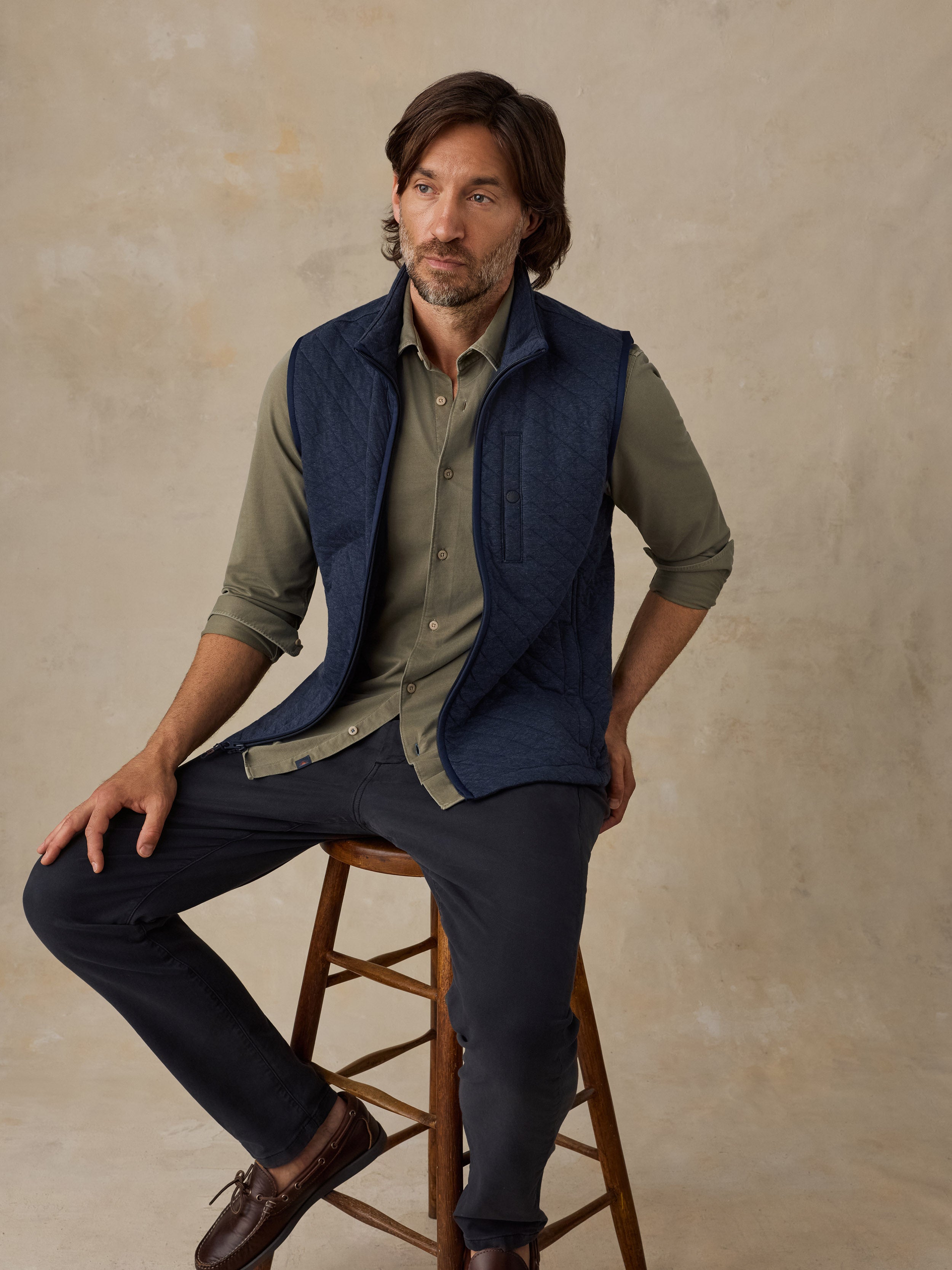 A man wearing a navy blue quilted vest over an olive green button-down shirt, dark trousers, and brown shoes, sitting on a wooden stool against a textured beige studio backdrop.