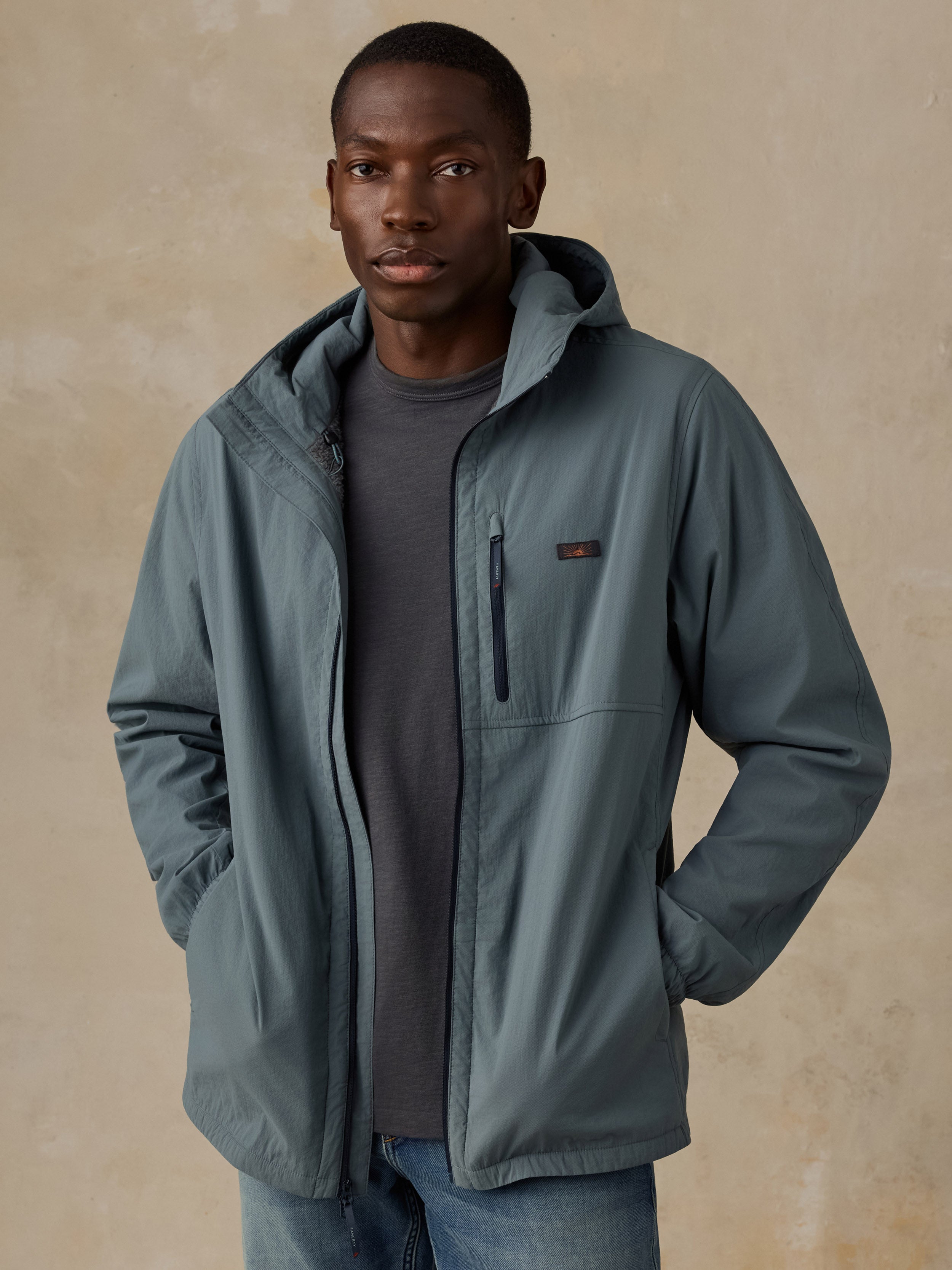 A man wearing a slate blue-gray hooded jacket over a dark gray t-shirt and blue jeans, standing against a textured beige background.