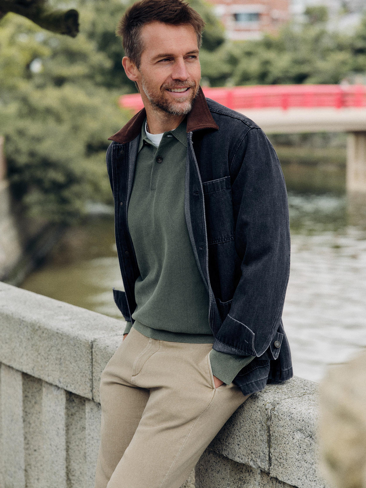 A man wearing a dark charcoal corduroy overshirt with a brown collar, an olive green polo shirt, and beige trousers, leaning against a stone bridge railing with water and a red bridge in the background., image 1 of 7