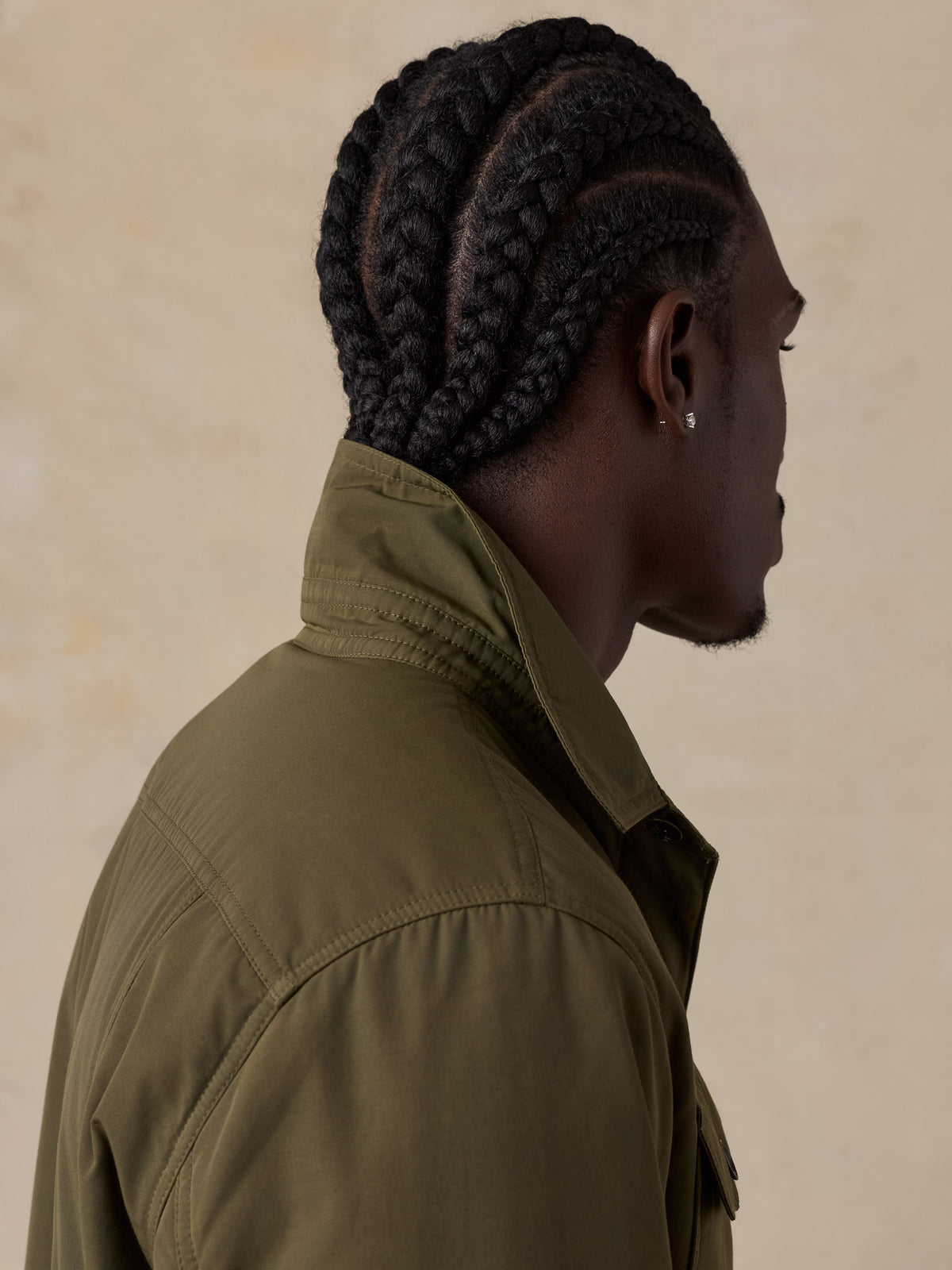 A man shown in profile dressed in an olive green jacket against a textured beige background., image 6 of 6