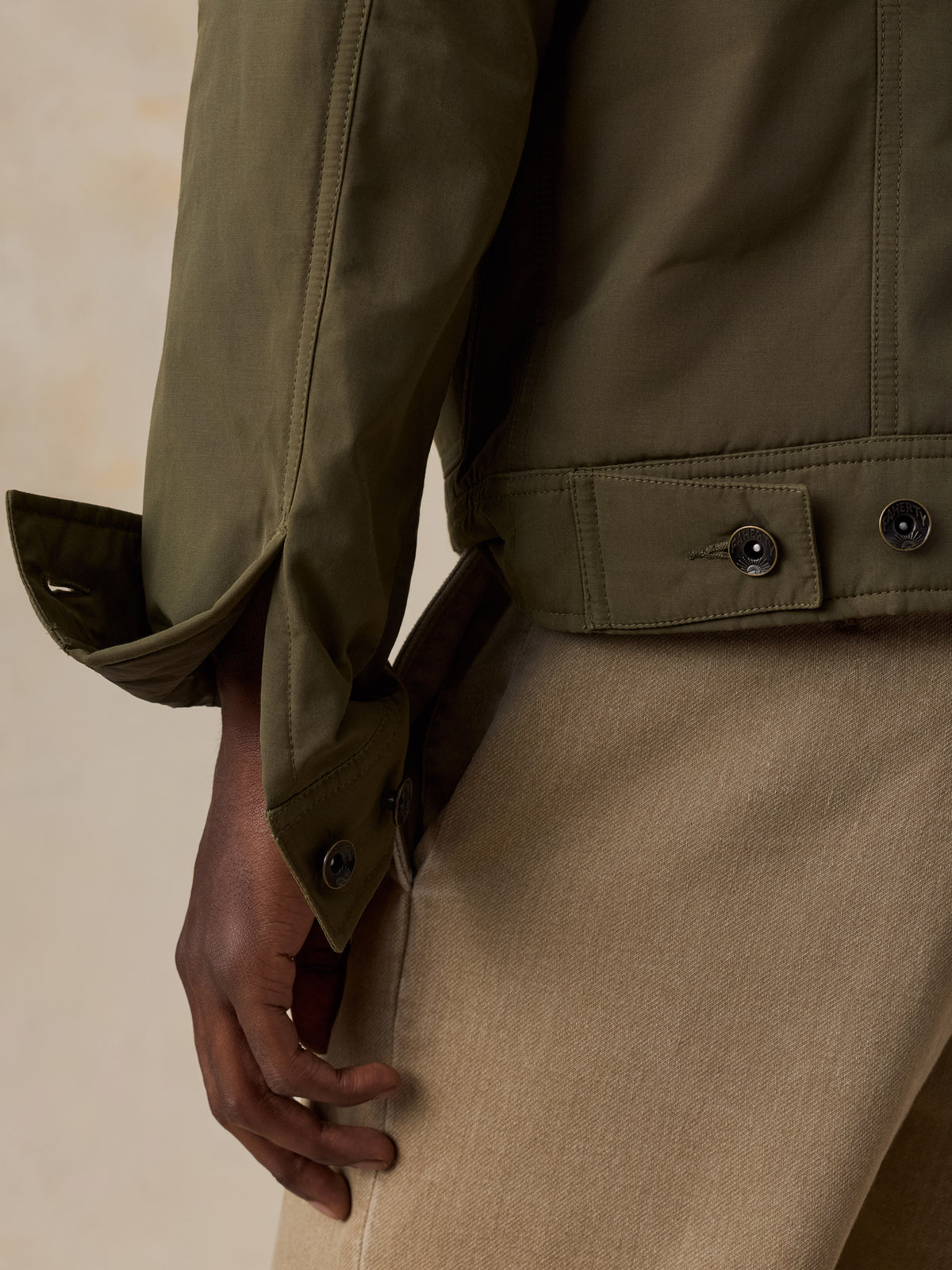 Close-up of a man wearing an olive green jacket with buttoned cuffs and a beige corduroy skirt against a warm beige background., image 5 of 6