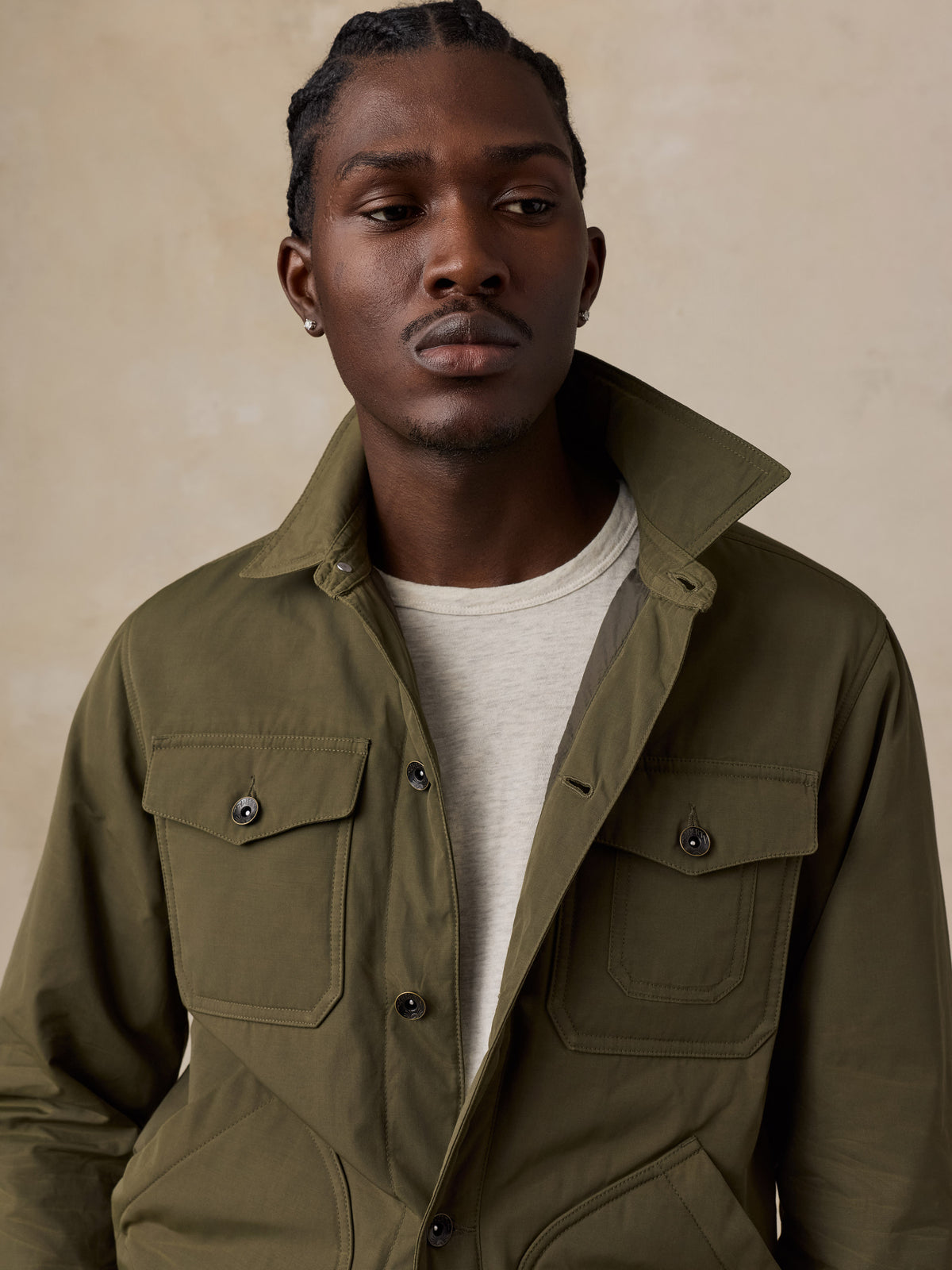 A man wearing an olive green utility jacket over a light beige t-shirt, photographed against a textured beige background., image 4 of 6