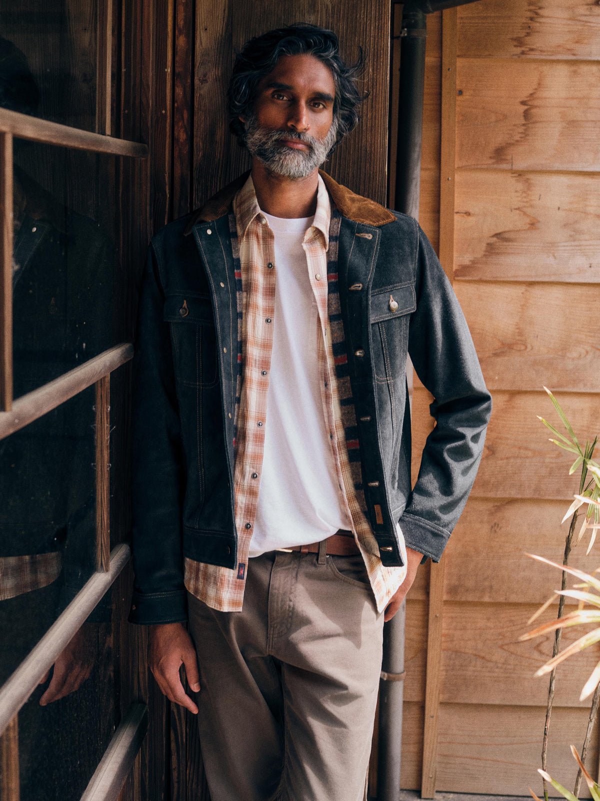 A man with a graying beard and dark hair stands in front of a rustic wooden door, wearing a dark charcoal suede jacket with brown collar lining over a beige and orange plaid flannel shirt, a white t-shirt, and khaki pants., image 1 of 8