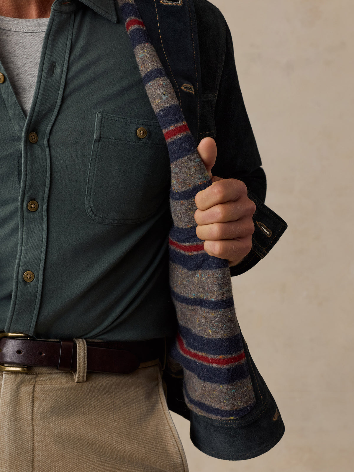 A close-up of a man wearing a dark green button-up shirt, beige trousers, and a dark overshirt, holding a striped wool sweater draped over their shoulder against a neutral beige background., image 4 of 8