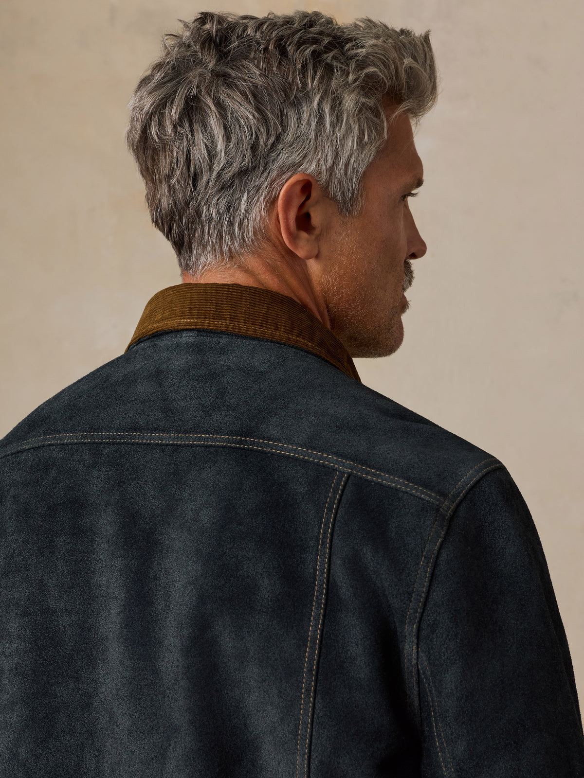 A man shown from the back and side profile wearing a dark charcoal gray suede jacket with a brown corduroy collar, set against a warm beige textured background., image 8 of 8
