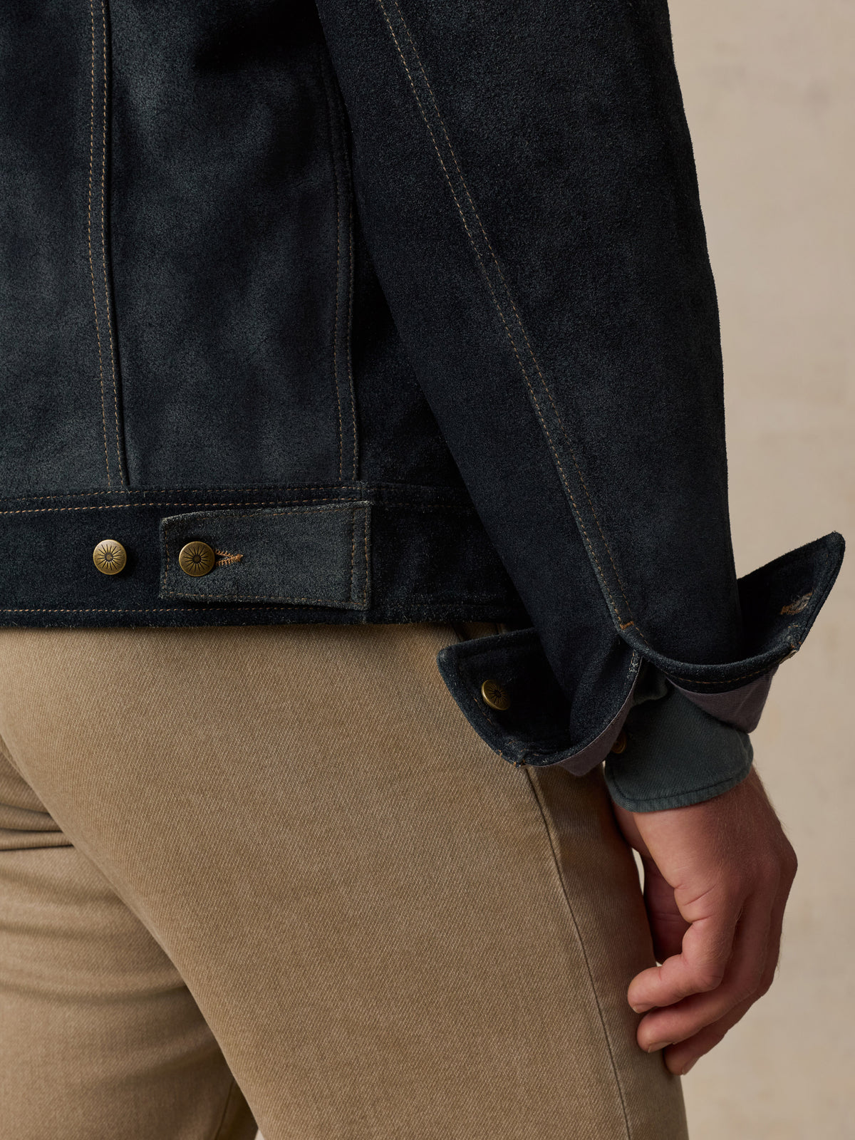 Close-up of a man wearing a dark indigo denim jacket with rolled cuffs and tan pants, showing the sleeve, button details, and hand against a neutral beige background., image 7 of 8