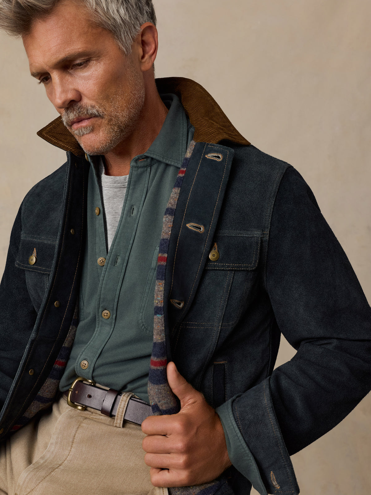 A close-up of a man wearing a dark navy waxed cotton jacket with a brown corduroy collar, layered over a sage green button-down shirt and beige trousers., image 6 of 8