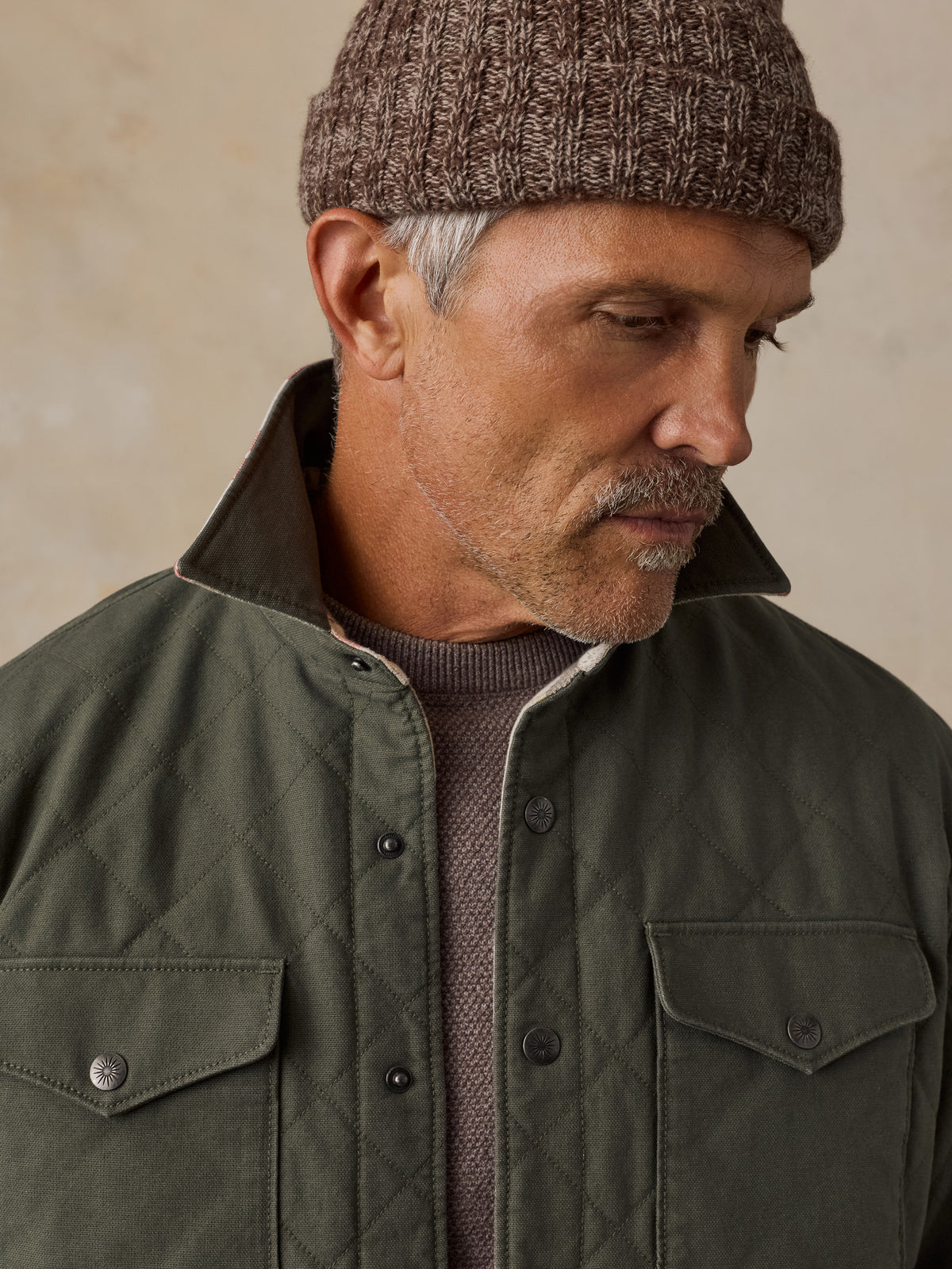 A close-up of a man wearing an olive green quilted jacket over a brown knit sweater and a brown knit beanie, set against a neutral beige background., image 5 of 5