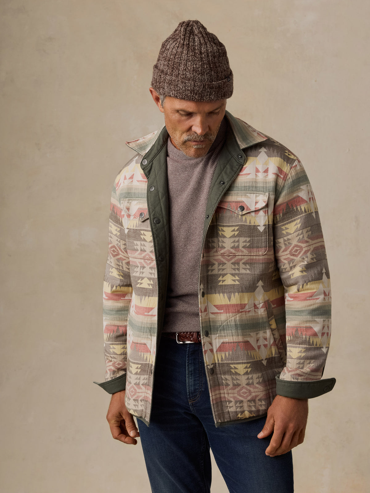A man wearing a patterned button-up shirt jacket with a Southwestern geometric design, layered over a gray sweater and blue jeans, paired with a brown knit beanie, standing against a neutral beige background., image 2 of 5