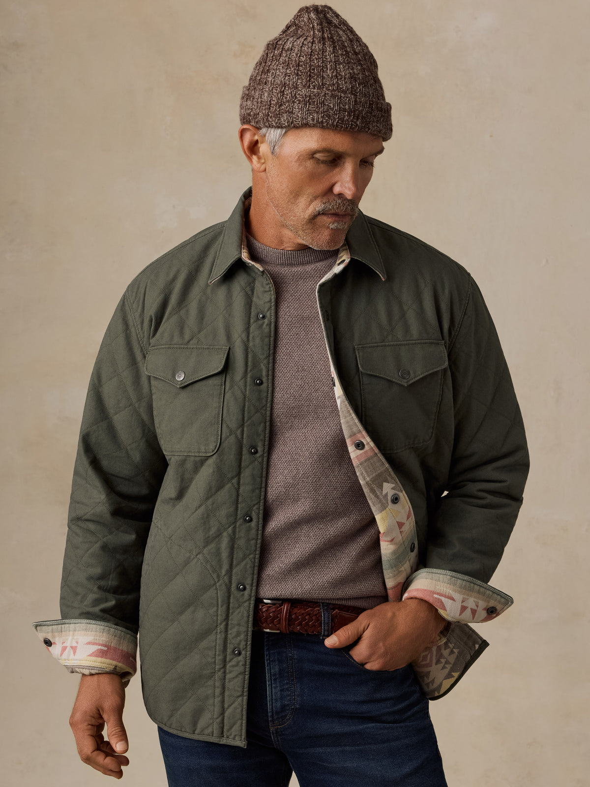 A man wearing an olive green quilted button-up jacket with a patterned lining, a brown knit beanie, a gray t-shirt, and blue jeans, standing against a textured beige background., image 1 of 5