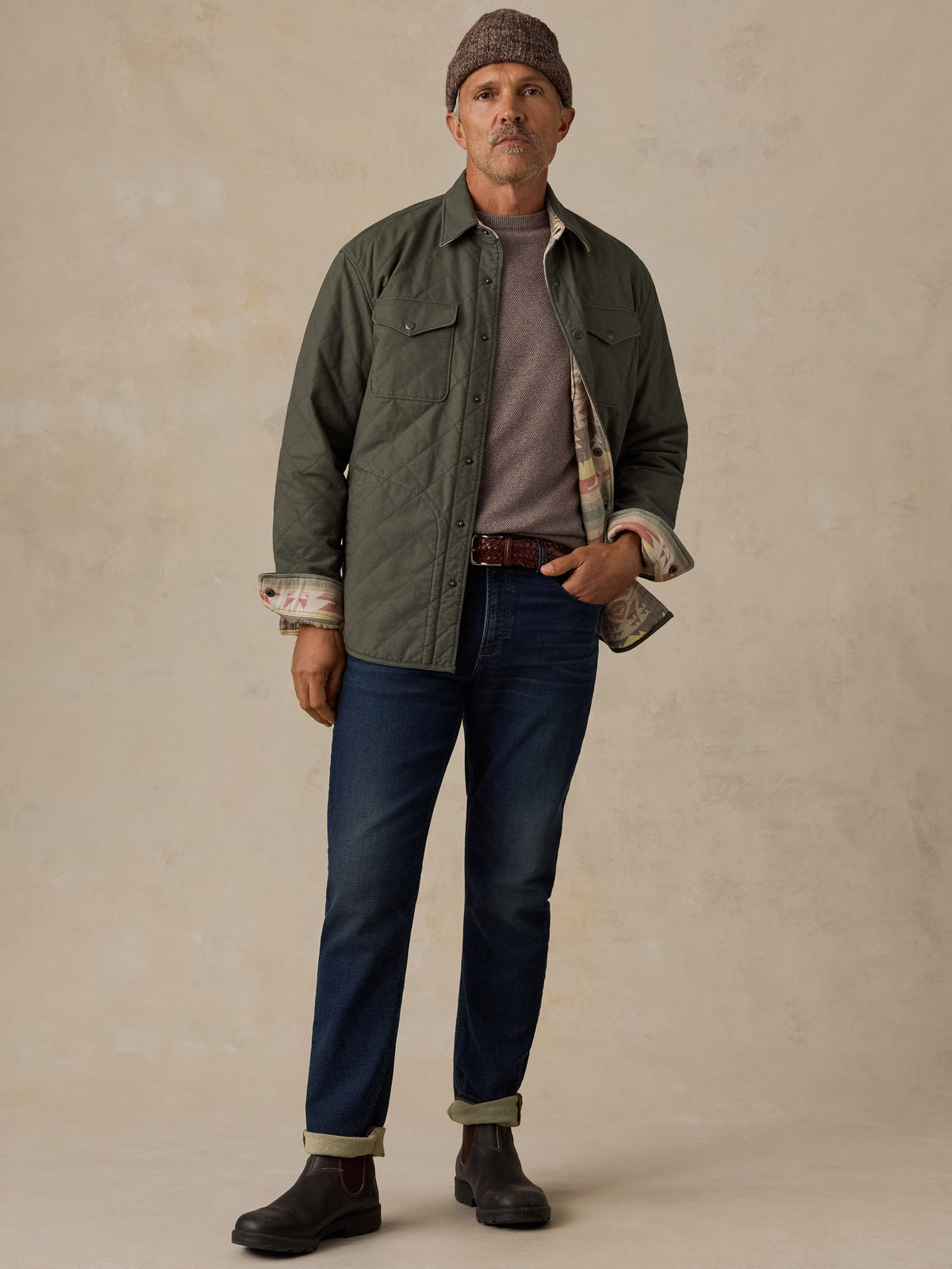 A man stands against a textured beige studio backdrop wearing a dark olive green quilted overshirt over a taupe knit sweater, blue jeans, brown boots, and a brown knit beanie., image 3 of 5