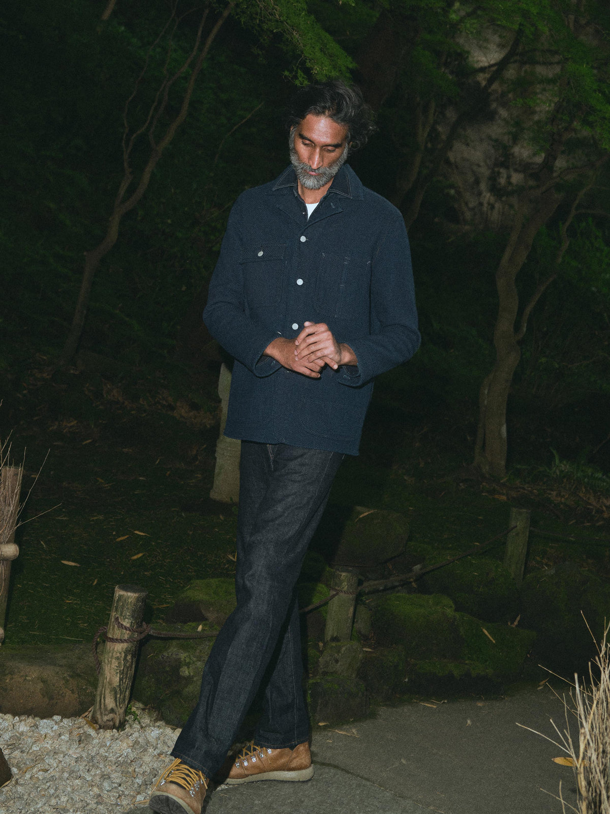 A man with a graying beard wearing a dark indigo denim button-up shirt over a white t-shirt and dark jeans, standing outdoors against a dark, blurry background of foliage., image 1 of 6