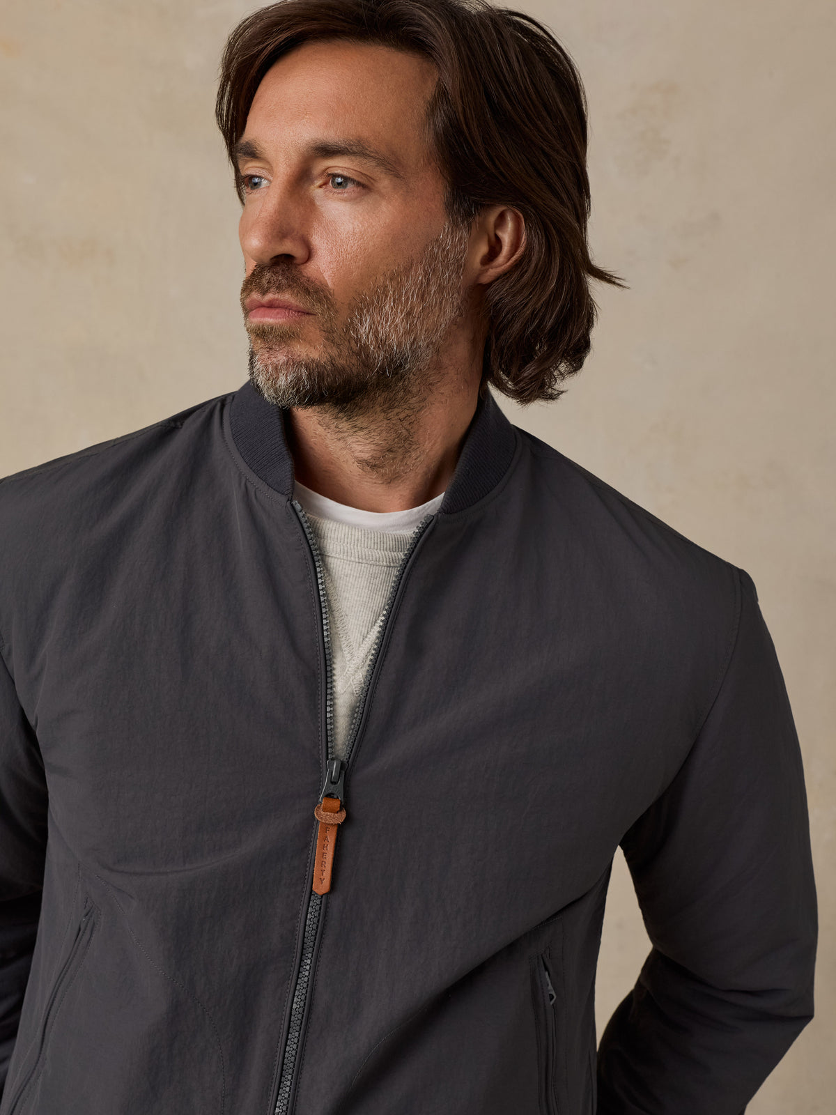 A man wearing a dark gray lightweight zip-up jacket over a light gray t-shirt, photographed against a textured beige background., image 5 of 6