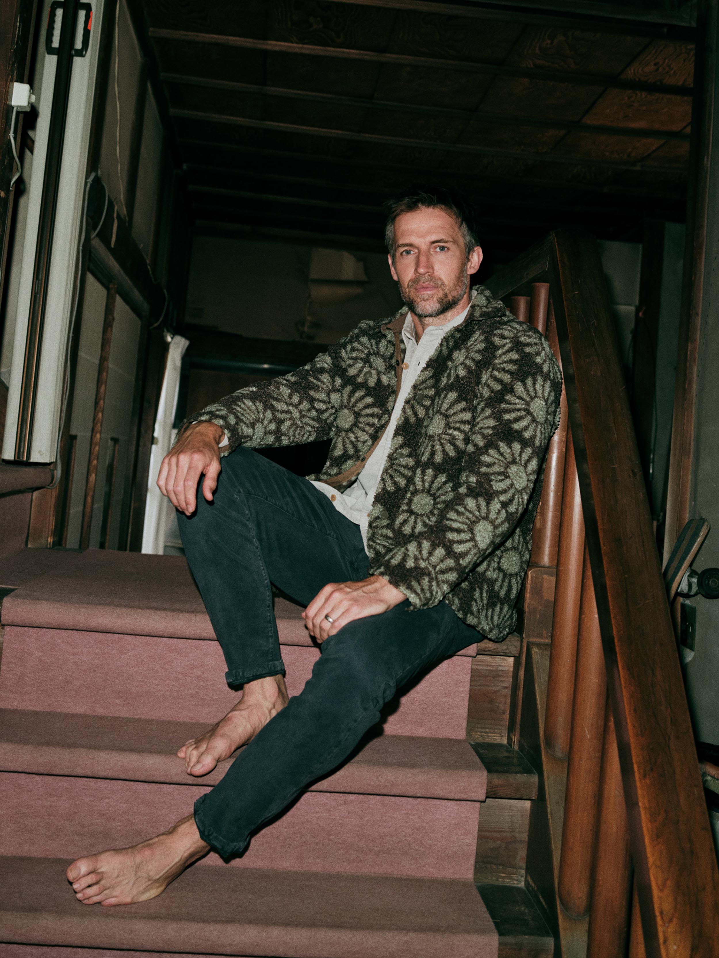A man sits on a wooden staircase indoors, wearing a floral patterned fleece jacket over a button up shirt and dark pants.