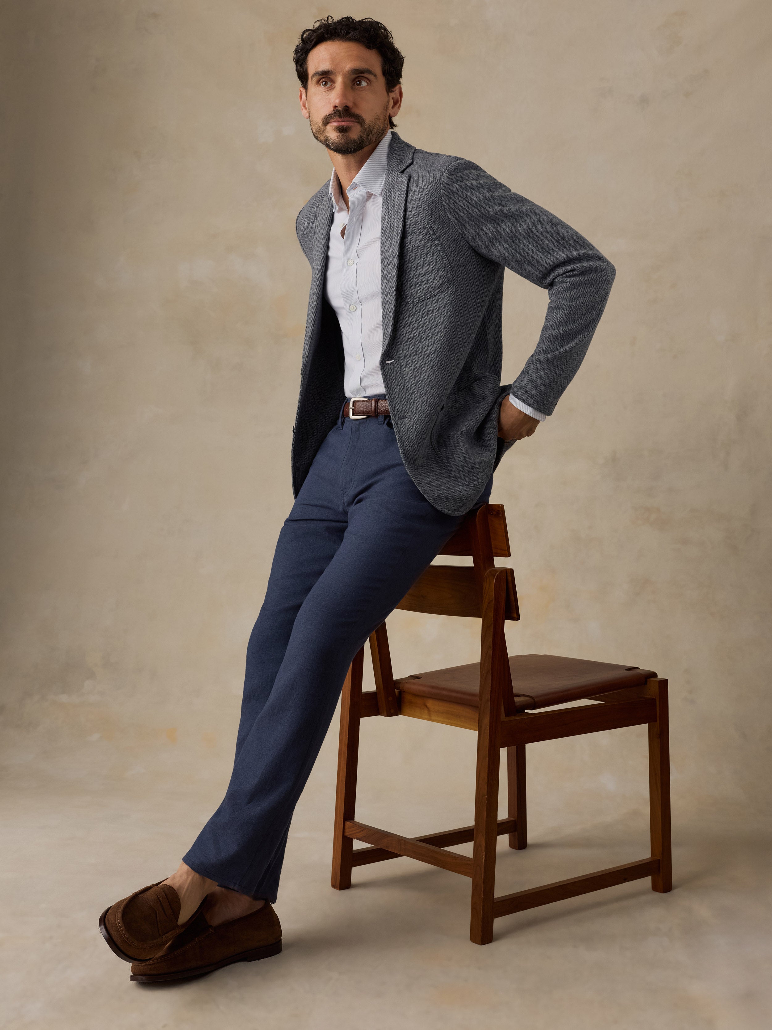 A man wearing a gray blazer, white shirt, navy trousers, and brown loafers leans against a wooden chair in a studio setting with a textured beige background.
