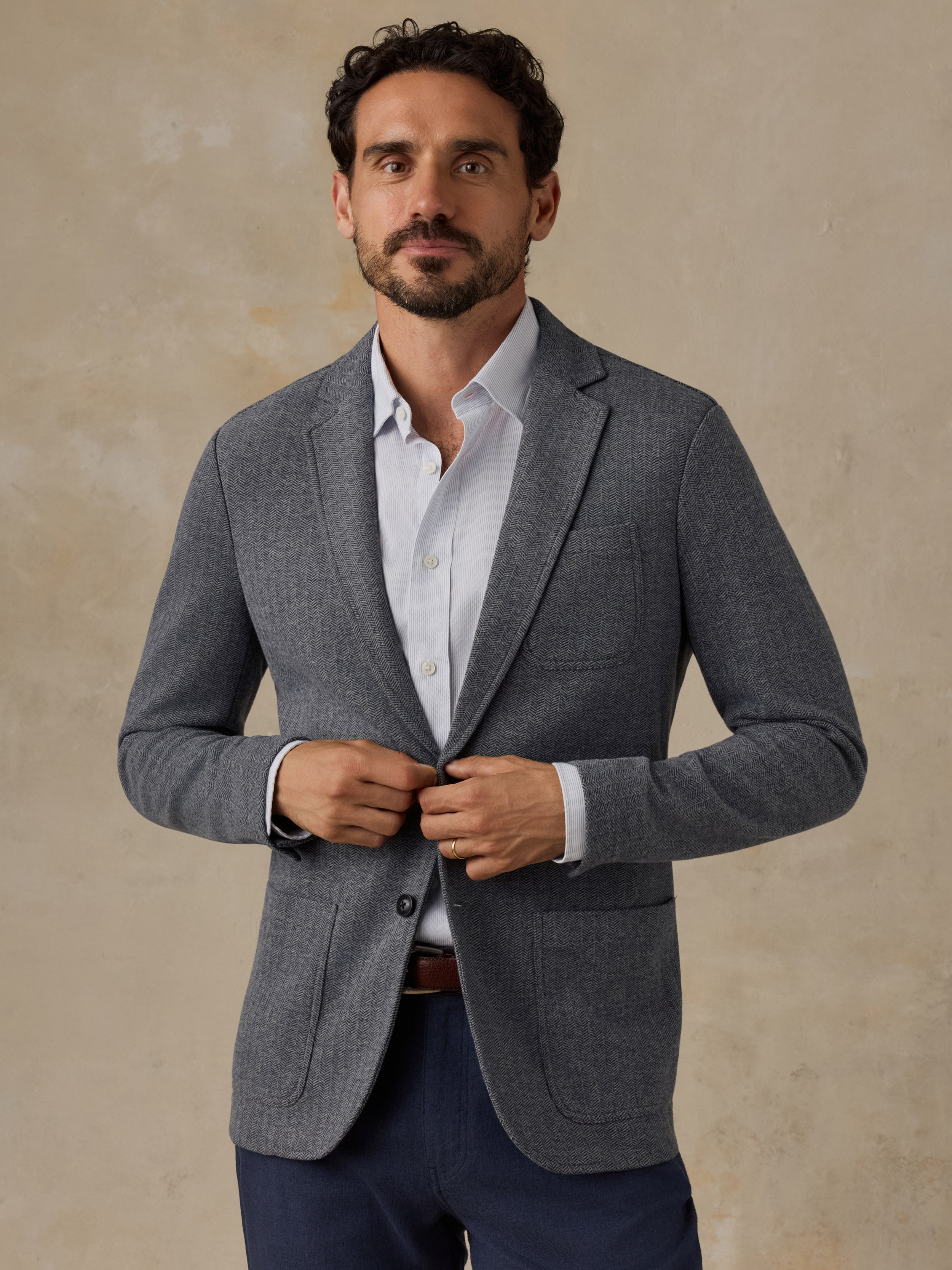 A man wearing a gray textured blazer over a white dress shirt and navy trousers, standing against a textured beige background.