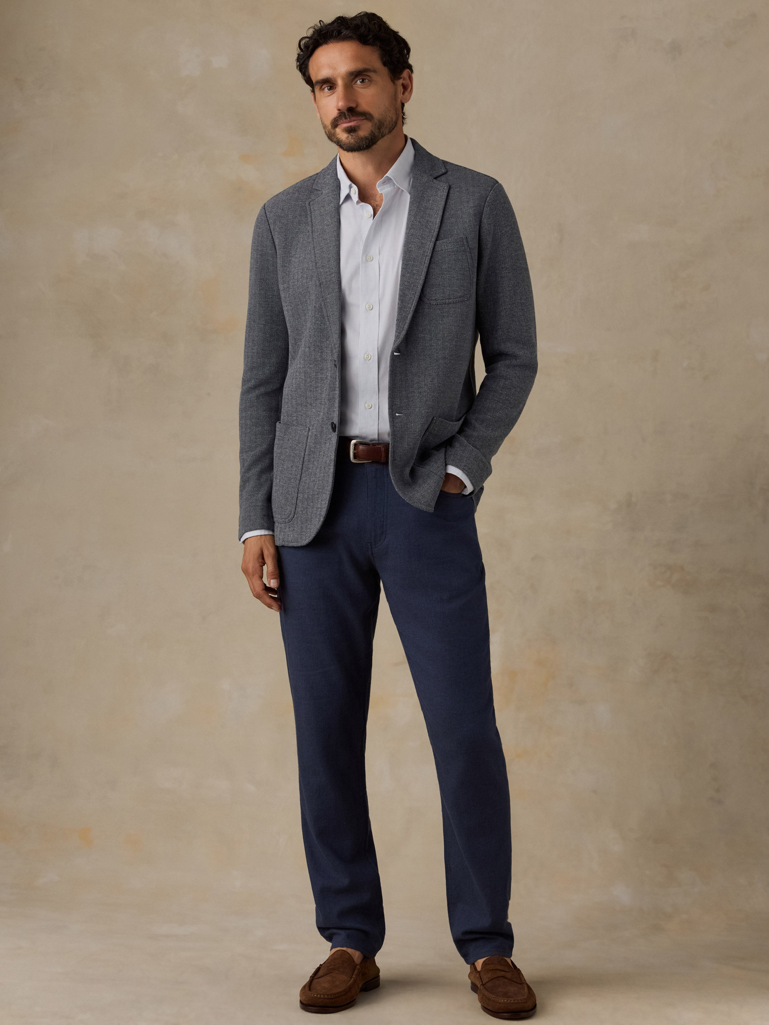A man wearing a gray textured blazer, white dress shirt, navy trousers, and brown suede loafers, standing against a textured beige studio backdrop.