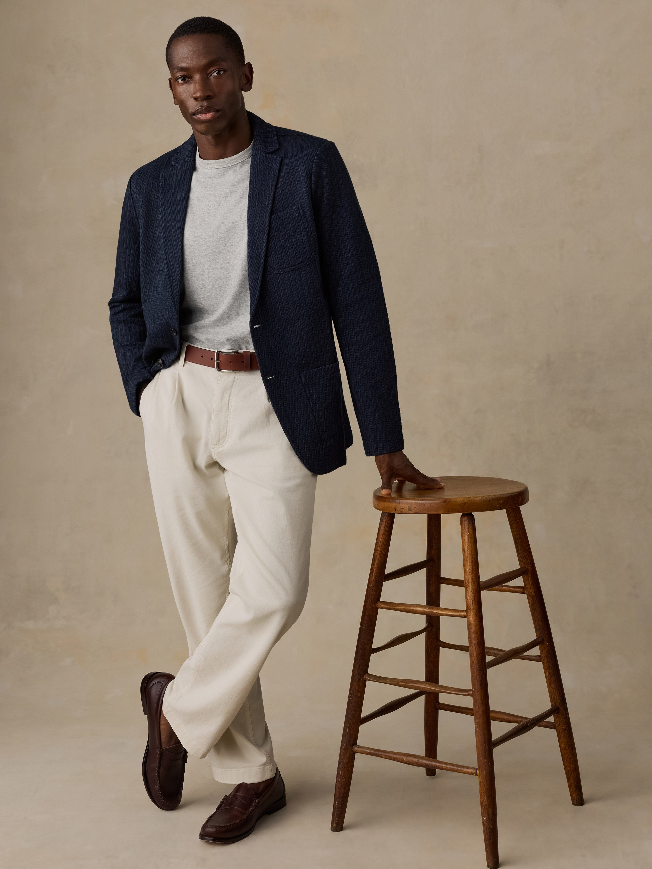 Man leaning on a wooden stool wearing a blue blazer over a grey t-shirt and white chinos against a beige background