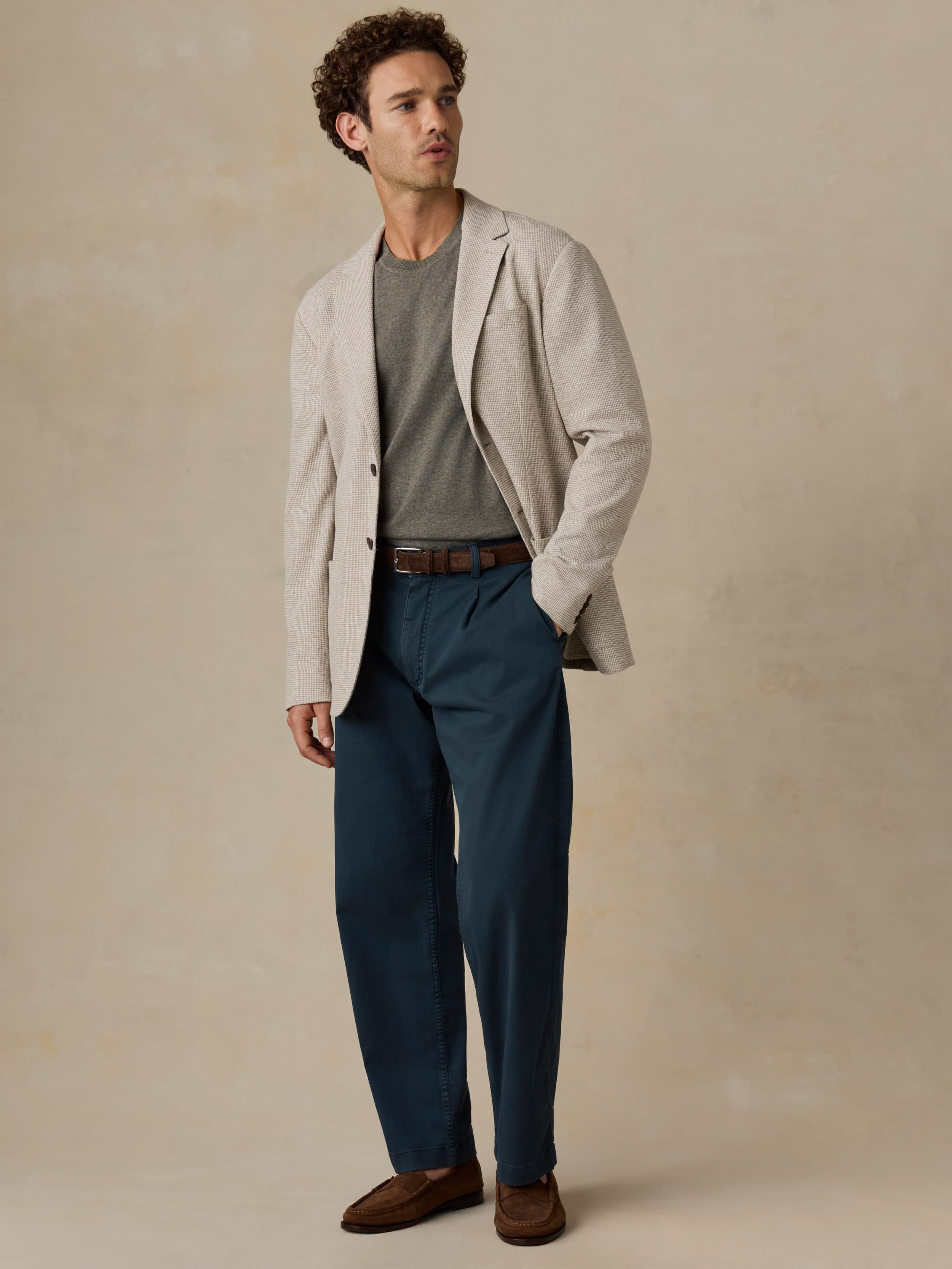Man wearing a beige blazer, gray shirt, blue pants, and brown shoes against a beige background