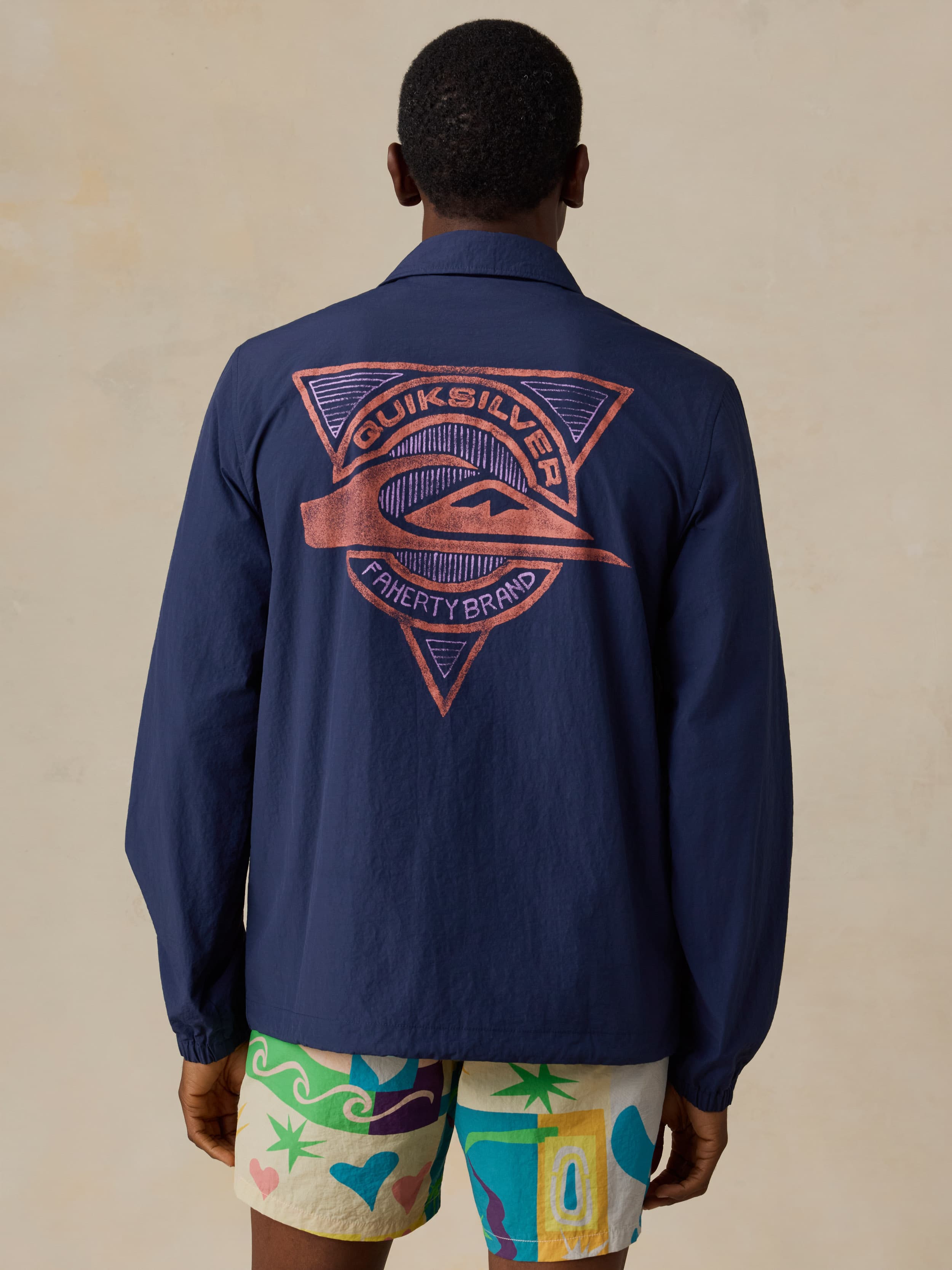 Man wearing a blue jacket with a Quiksilver x Faherty brand logo and colorful swim shorts against a beige background