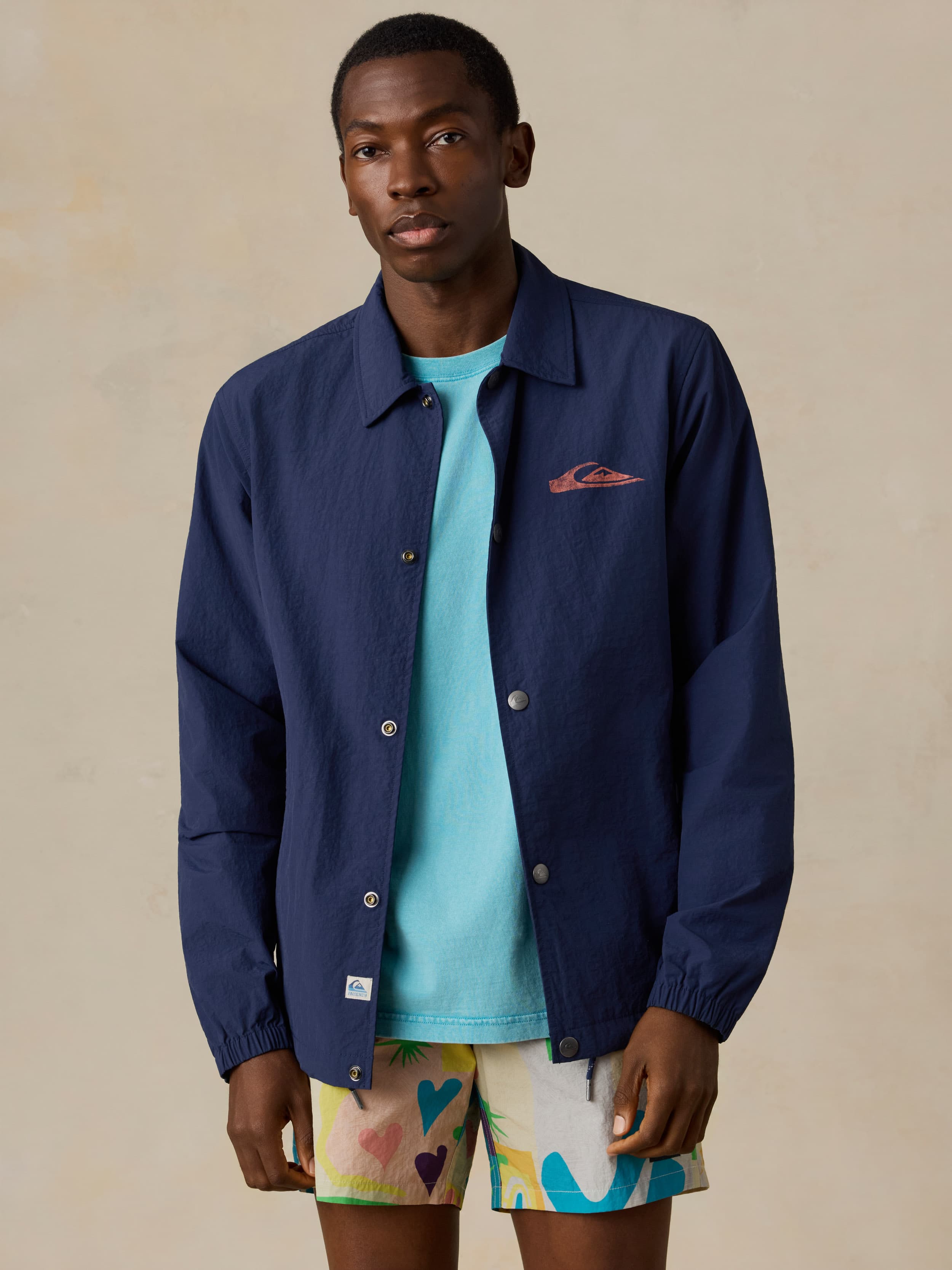 Man wearing a blue jacket with a Quiksilver x Faherty brand logo and colorful swim shorts against a beige background