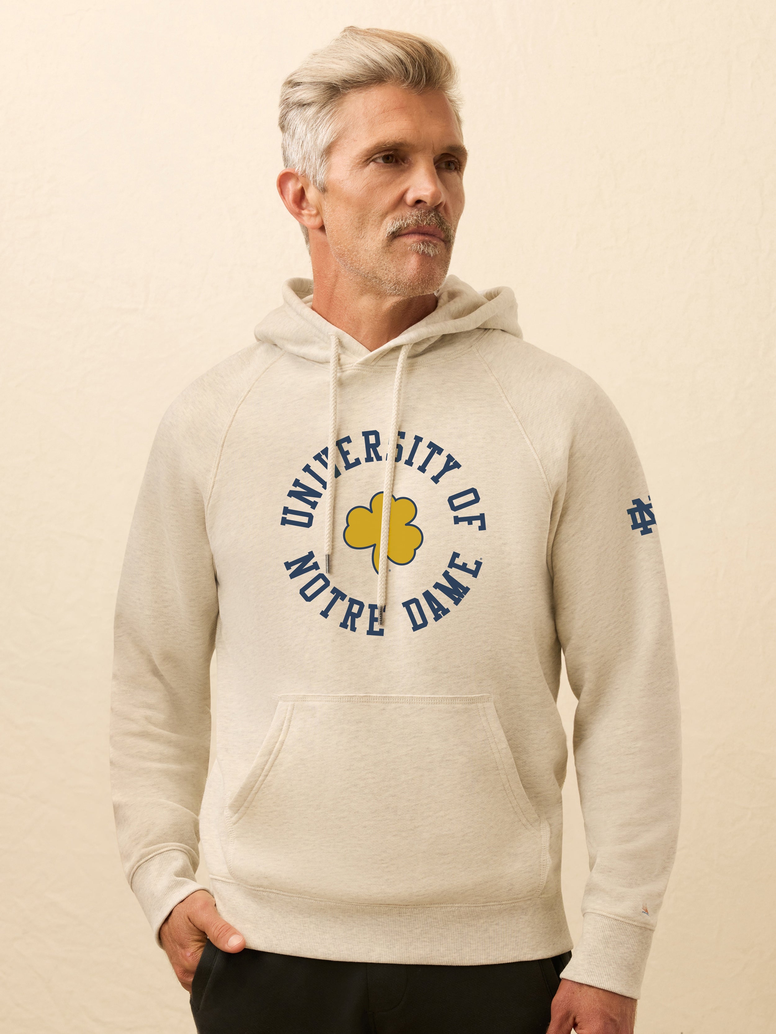 Man is wearing a heather grey hoodie with Notre Dame graphic on the center front against a beige background