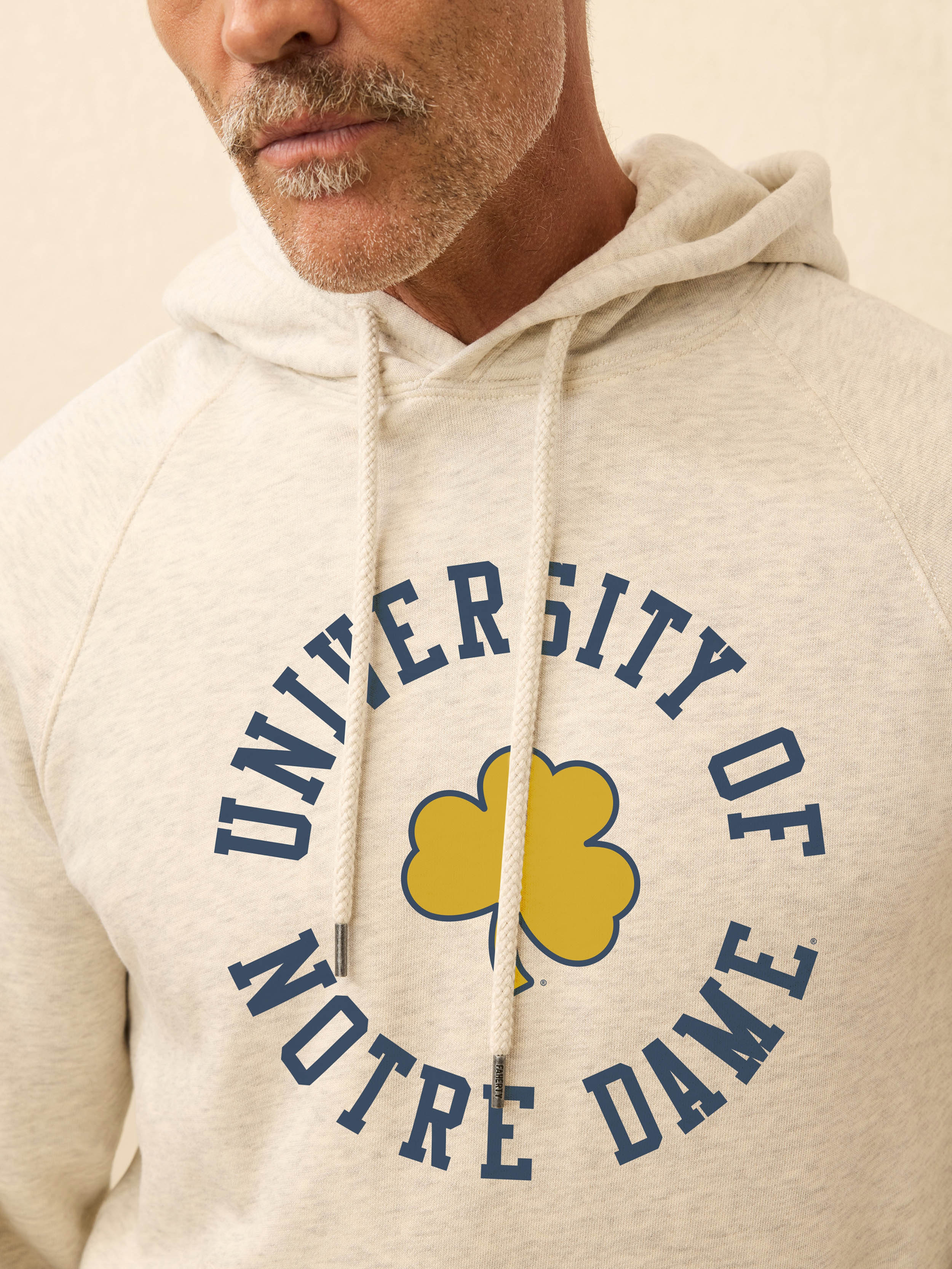 Man is wearing a heather grey hoodie with Notre Dame graphic on the center front against a beige background