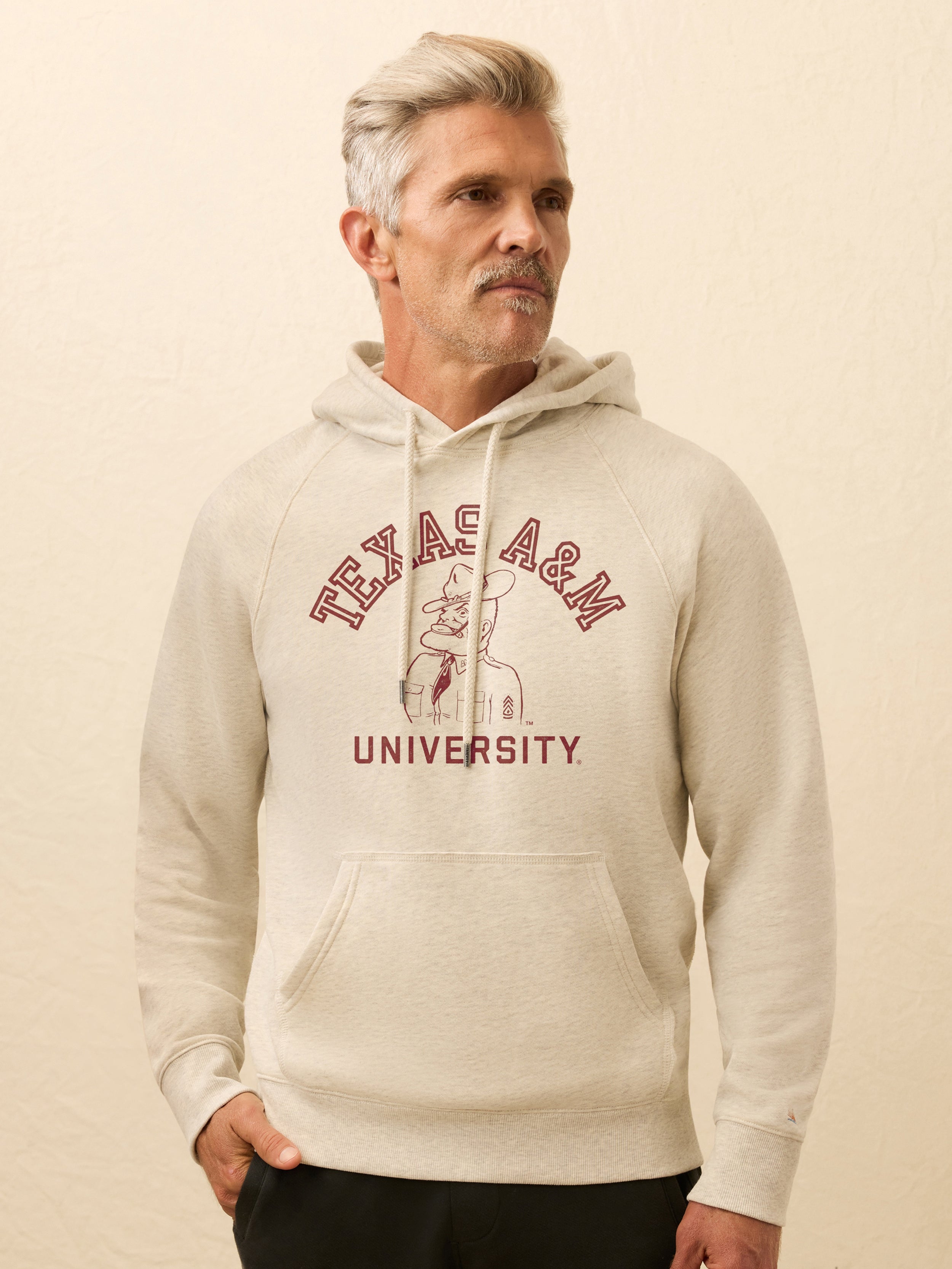 Man wearing an ivory hooded sweatshirt with Texas A&M graphic at the front against a beige background