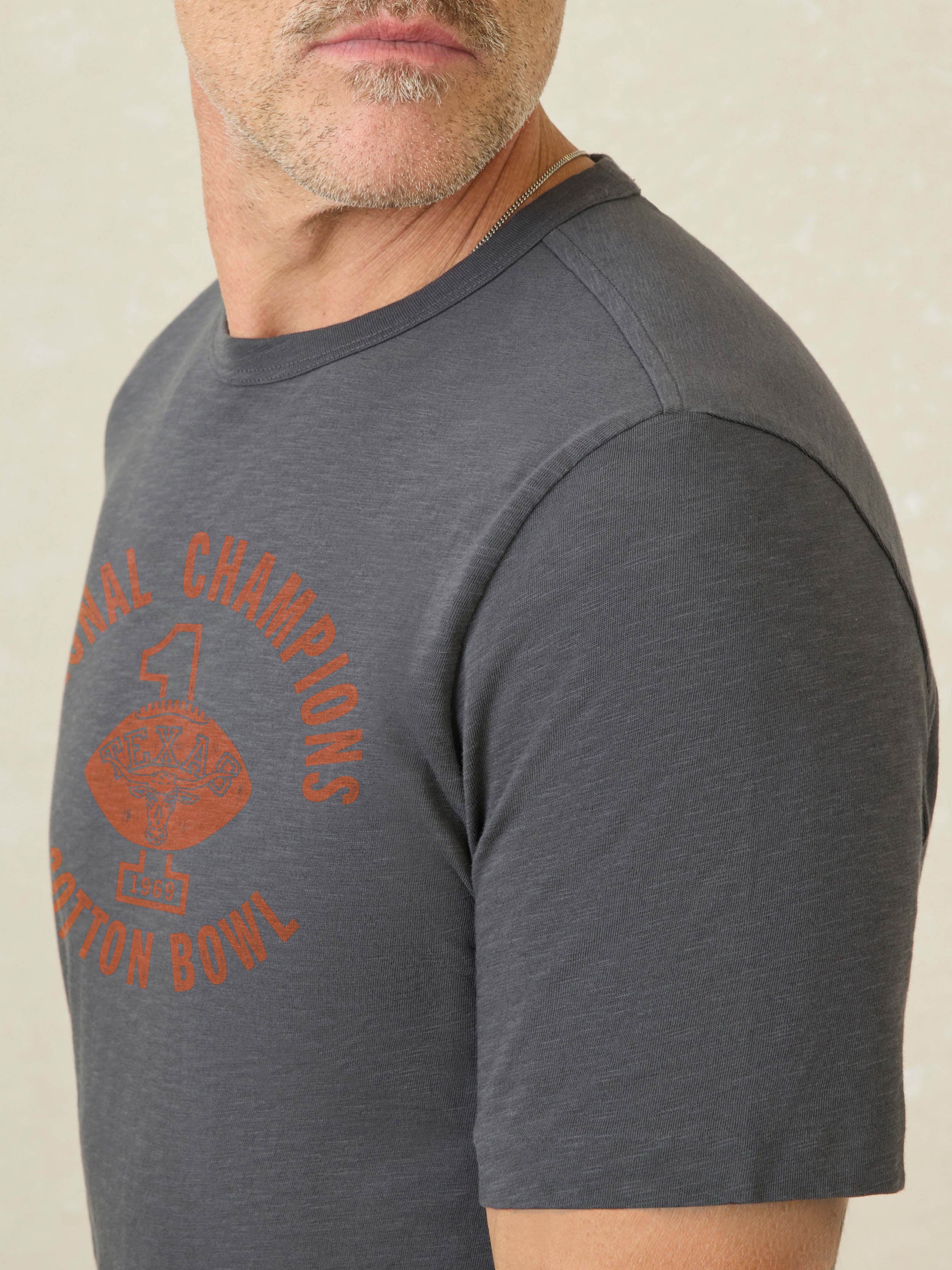 Man wearing a grey short sleeve t-shirt with UT Austin graphic on center front against a beige background