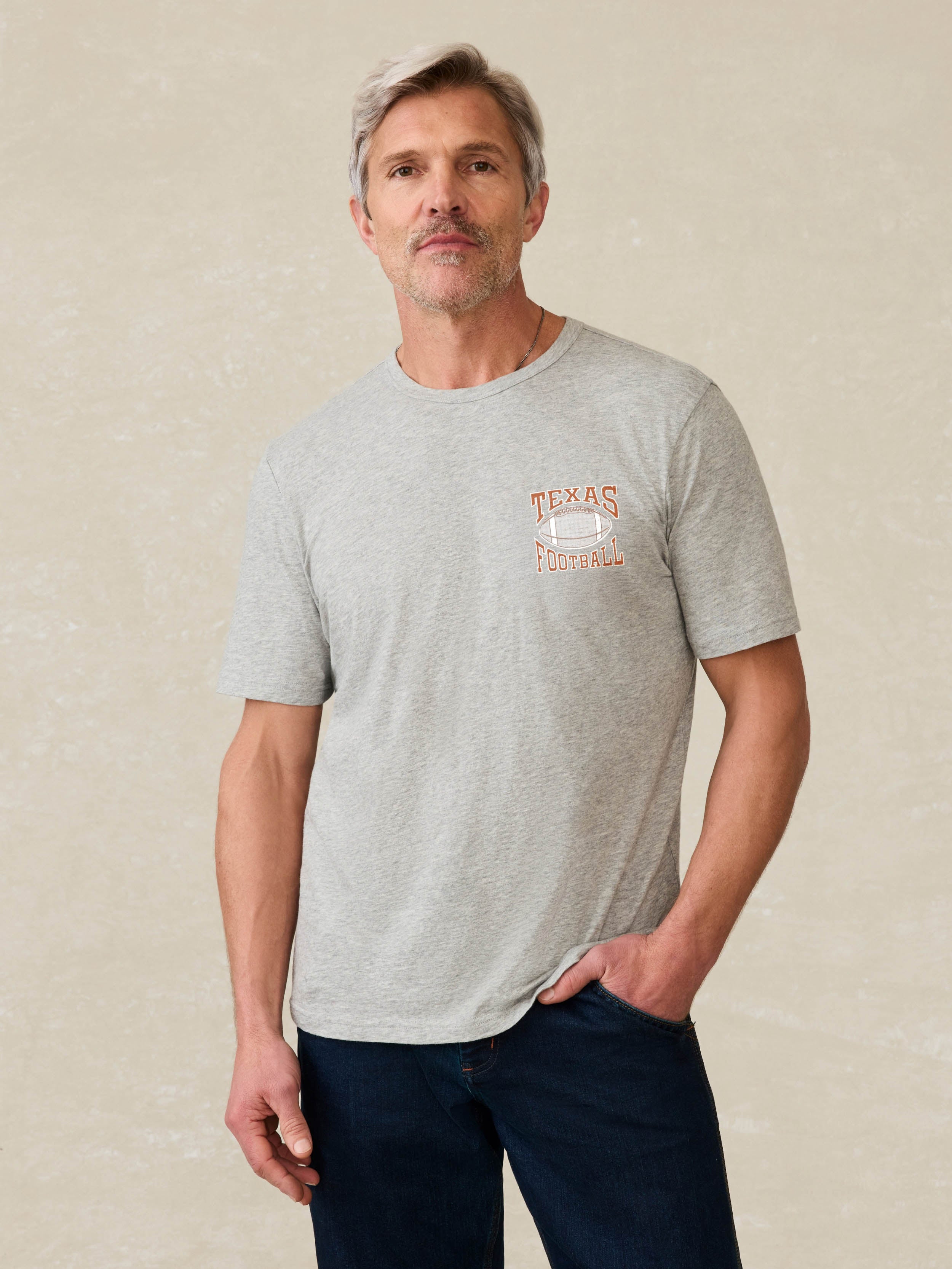 Man wearing a grey short sleeve t-shirt with UT Austin graphic on the left chest against a beige background