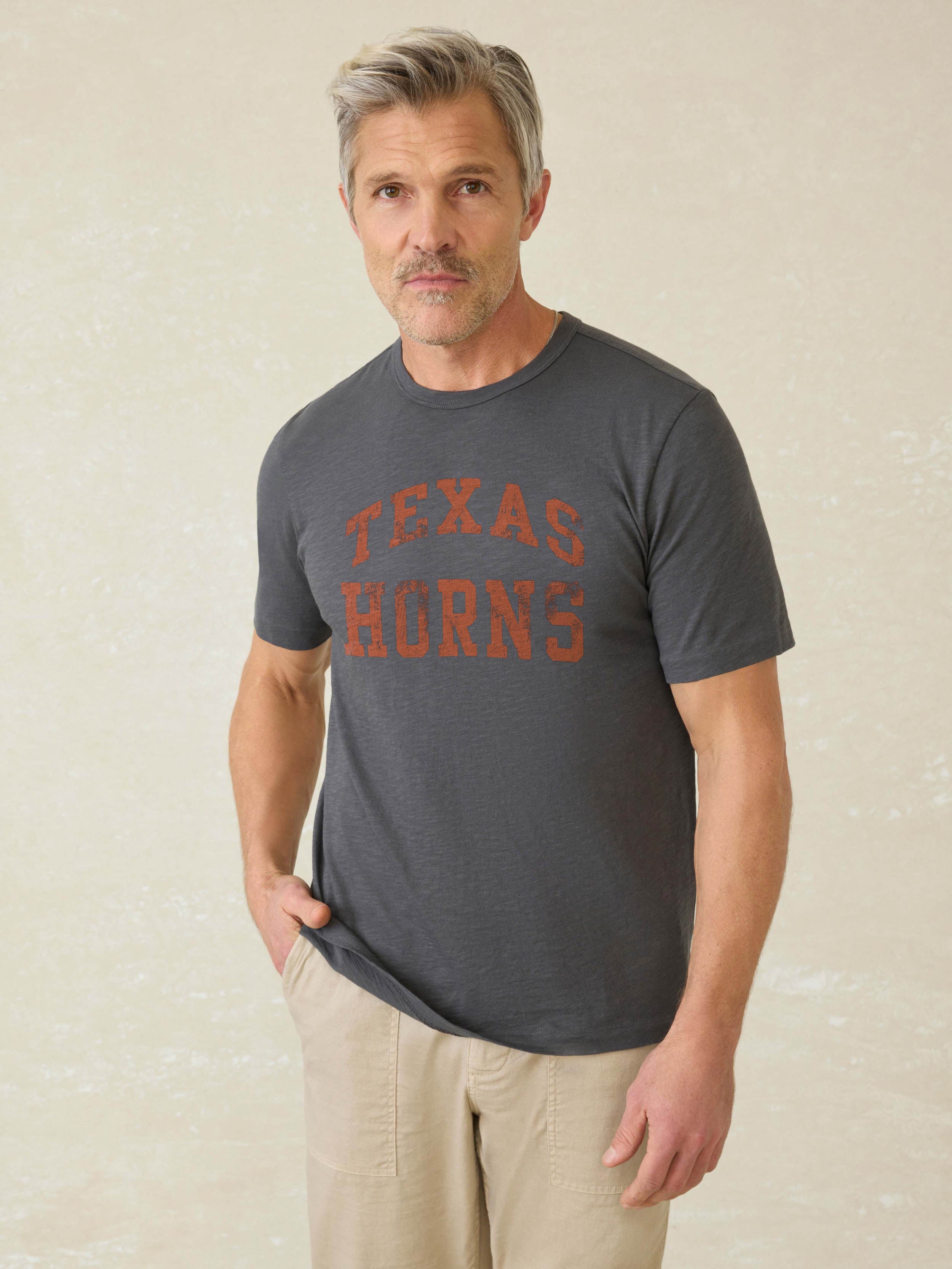 Man wearing a grey short sleeve t-shirt with UT Austin graphic logo on the center front against a beige background