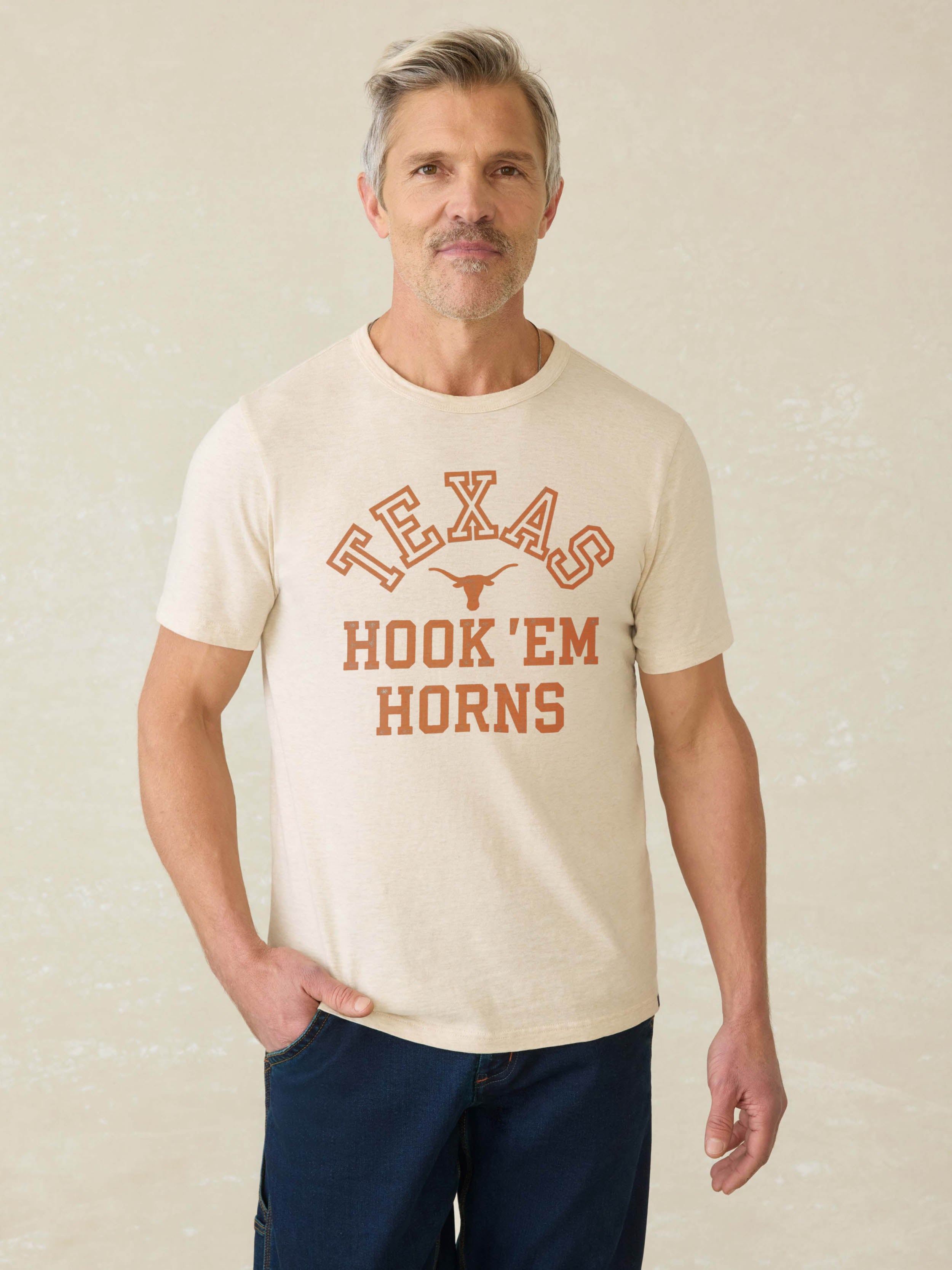 Man wearing a light grey short sleeeve t-shirt with UT Austing graphic on the center front against a beige background