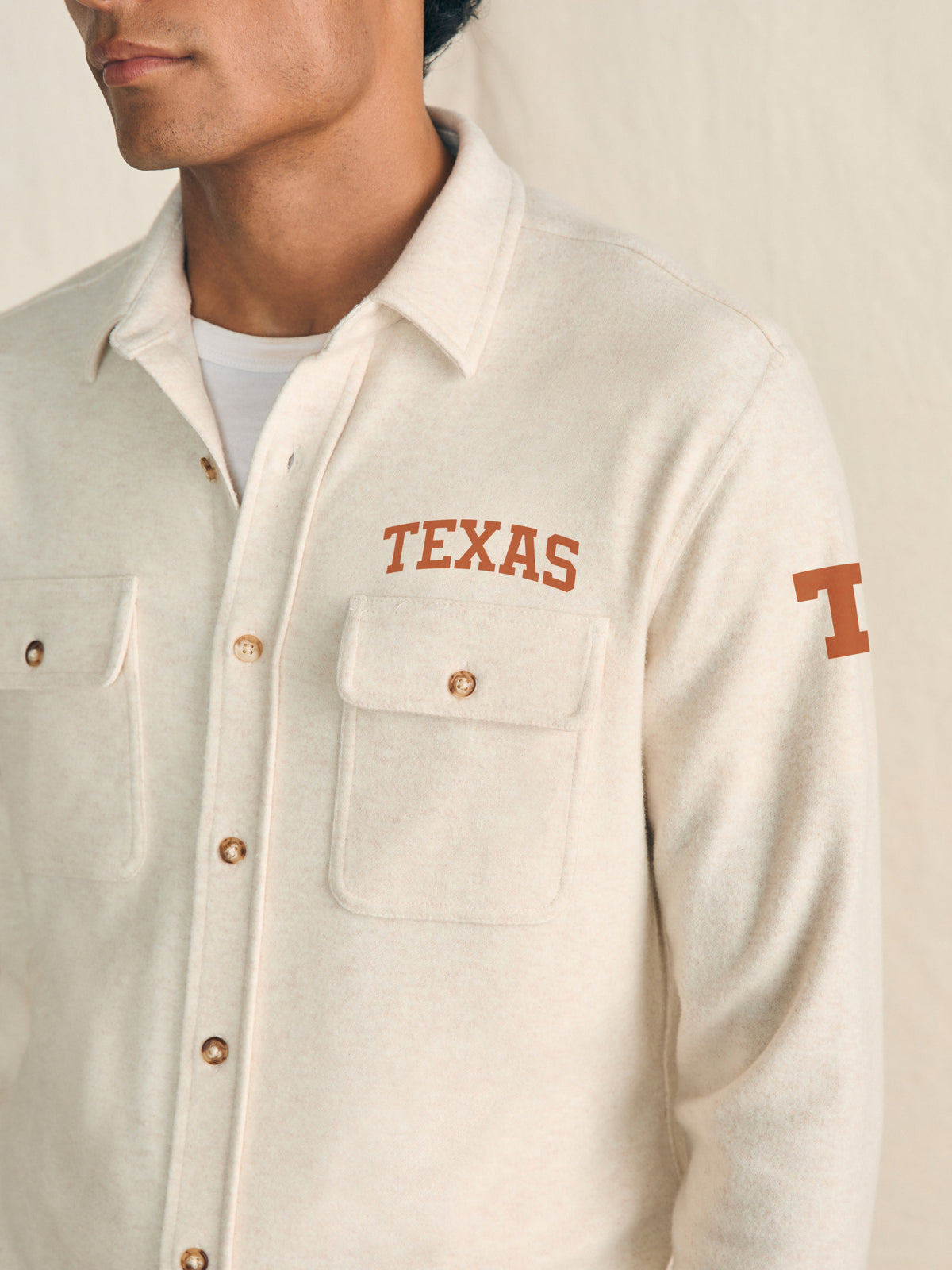 Man wearing a white shirt with UT Austin graphic on the left chest and shoulder against a beige background, image 2 of 3