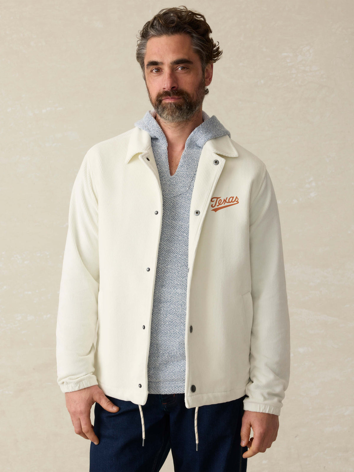 Man is wearing a white coaches jacket with UT Austin graphic on the left chest against a beige background, image 1 of 4