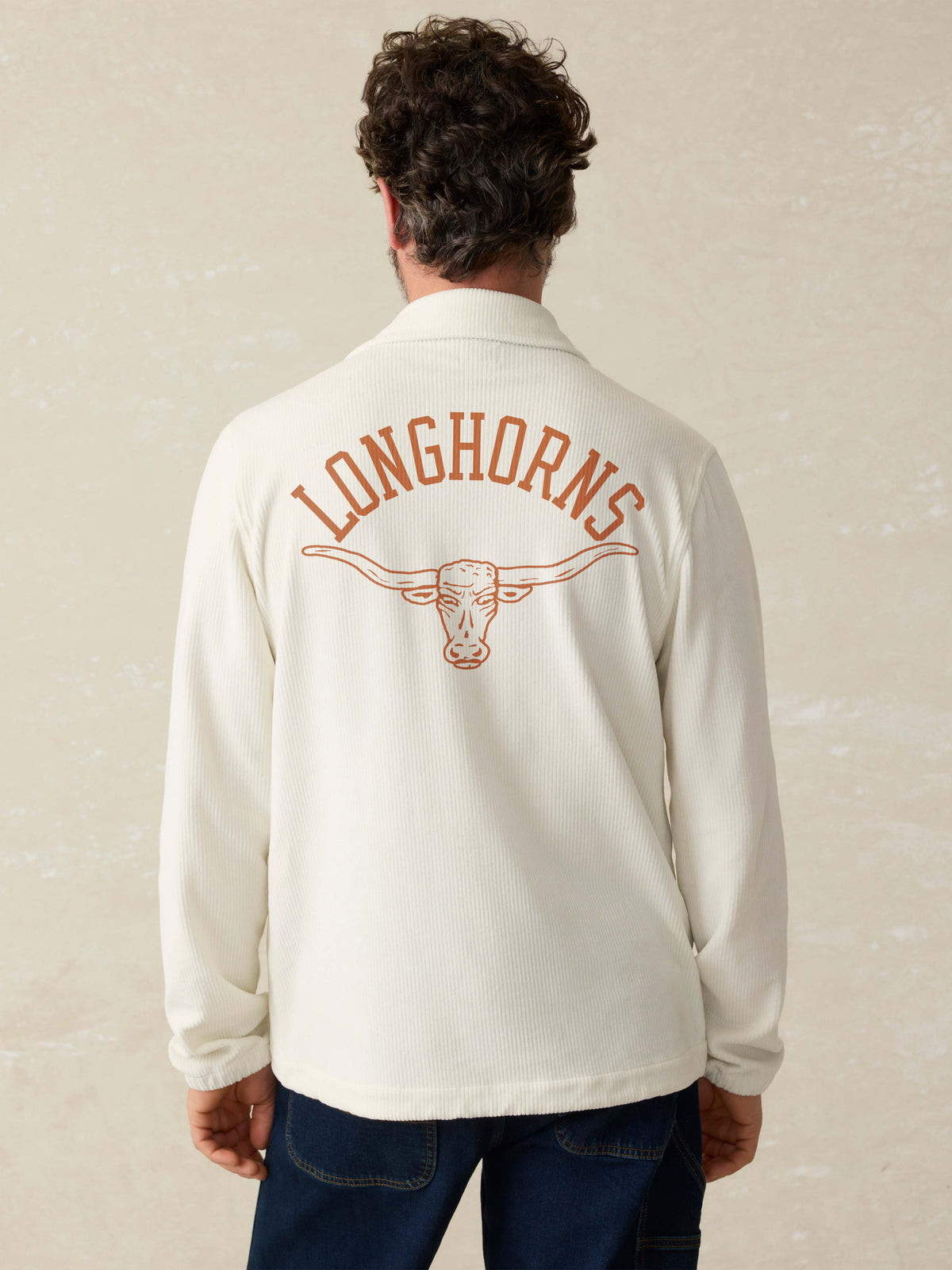 Man is wearing a white coaches jacket with UT Austin graphic on the center back against a beige background, image 2 of 4