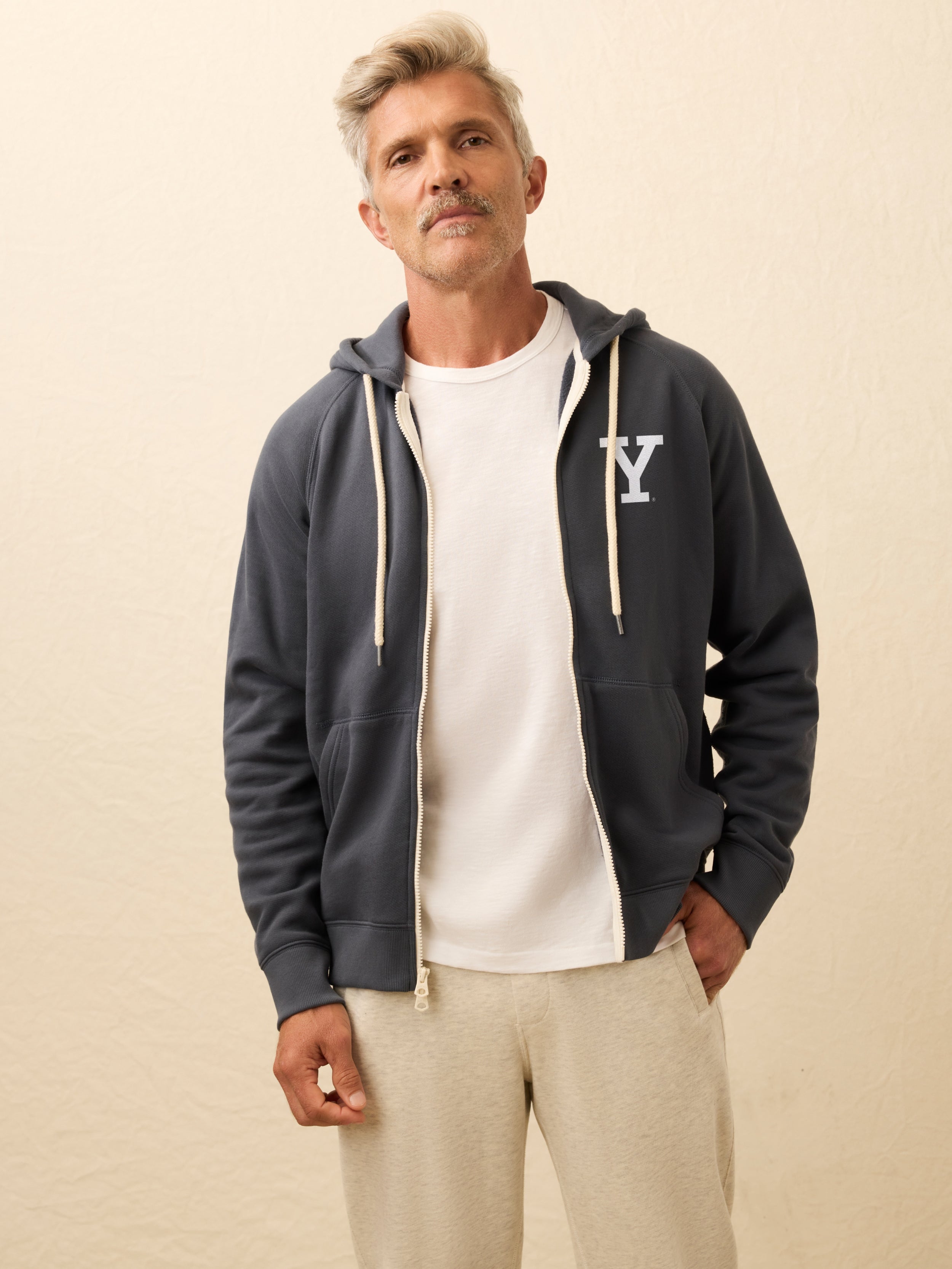 Man is wearing a navy zip hoodie with Yale graphic at left chest against a beige background