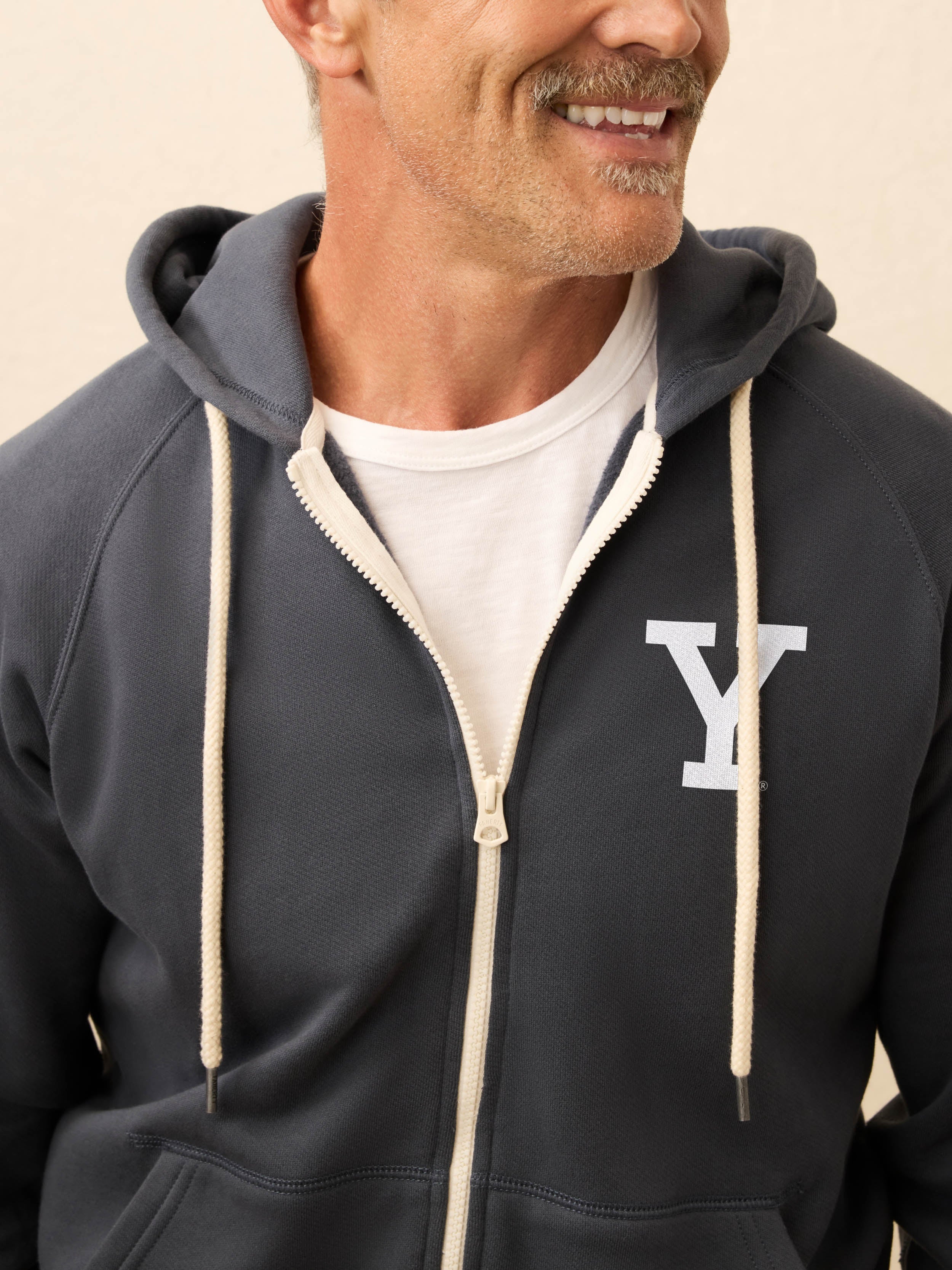 Man is wearing a navy zip hoodie with Yale graphic at left chest against a beige background