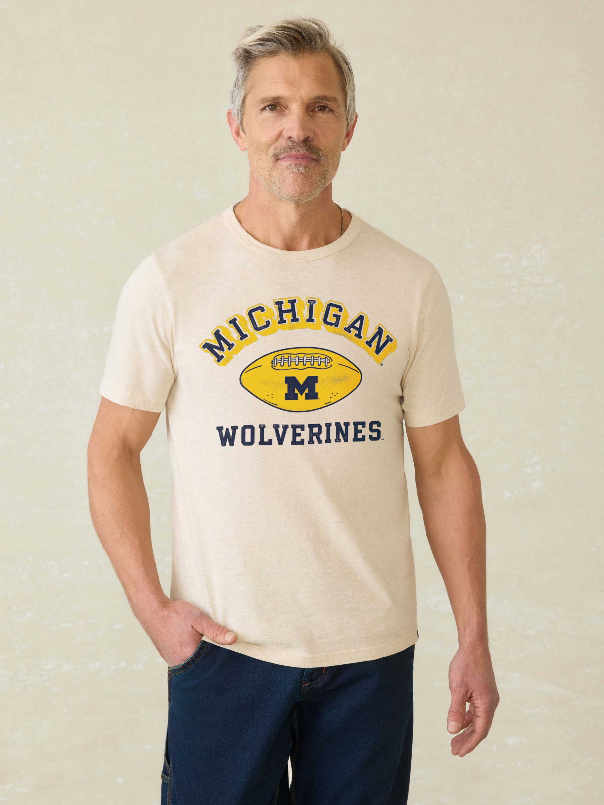 Man wearing a grey short sleeve t-shirt with University of Michigan graphic at the center chest against a beige background