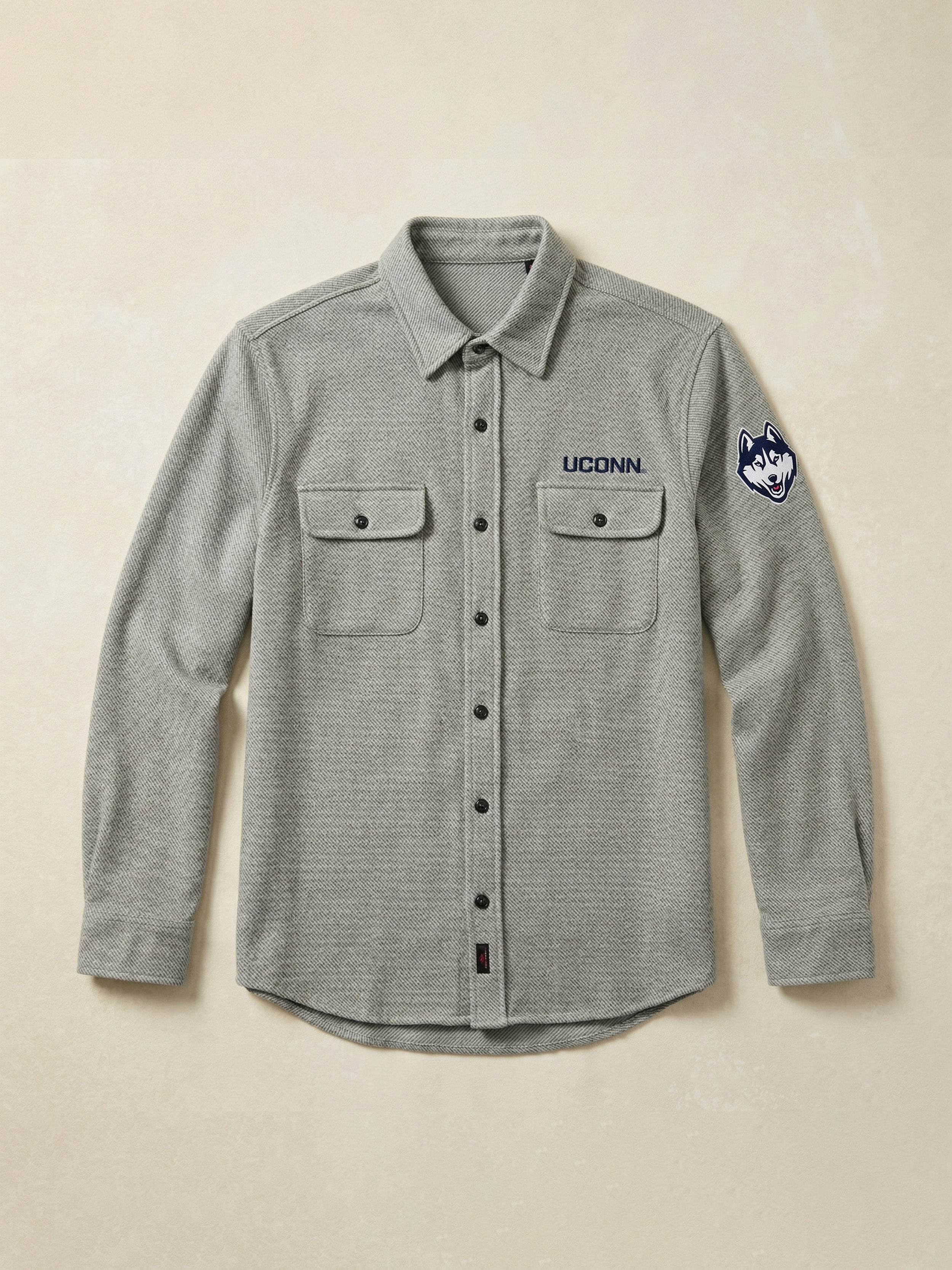 Gray flannel shirt with UConn logo on the left chest and the left shoulder against a beige background