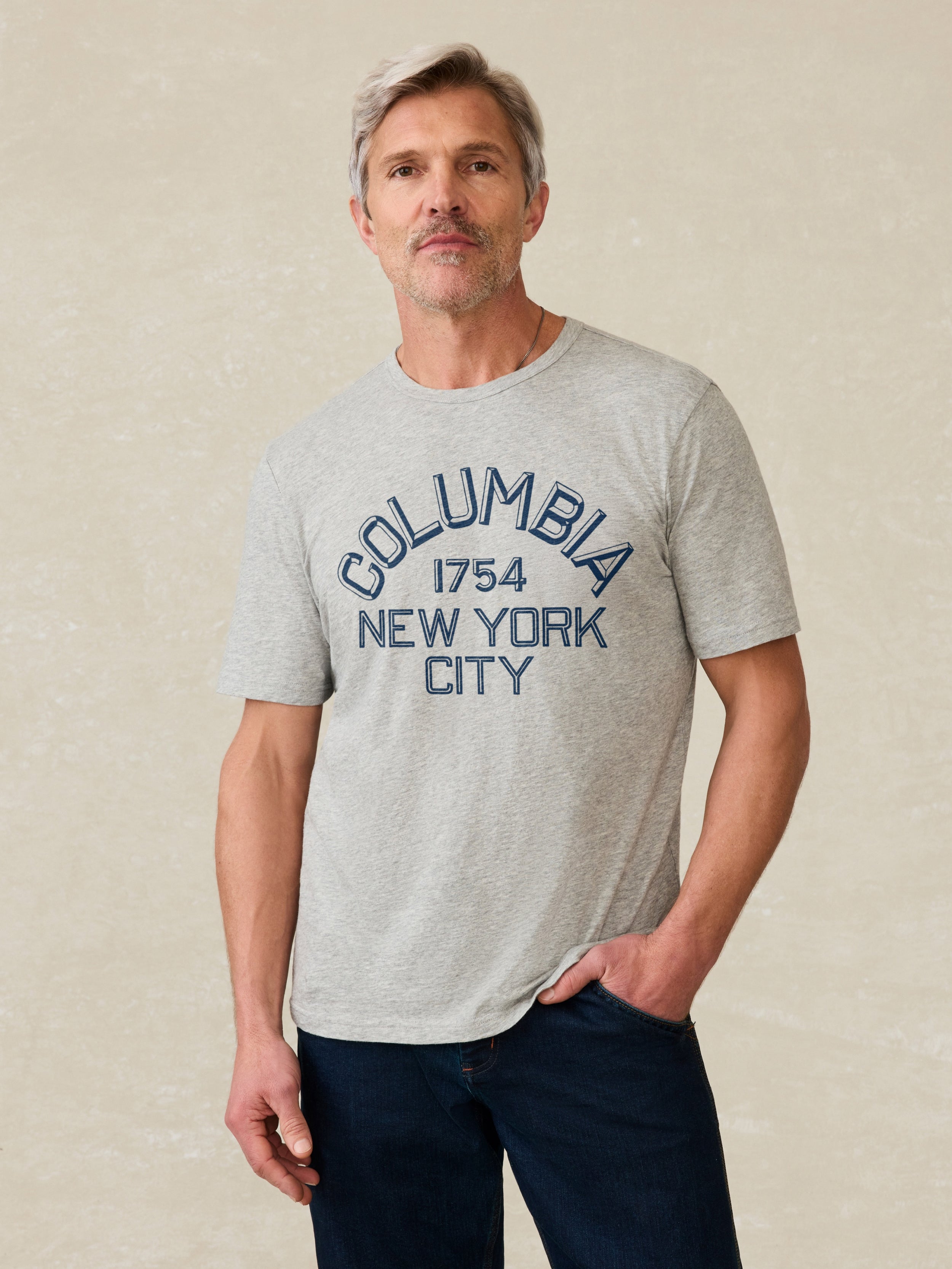 Man wearing a grey short sleeve t-shirt with Columbia graphic on the center front against a beige background