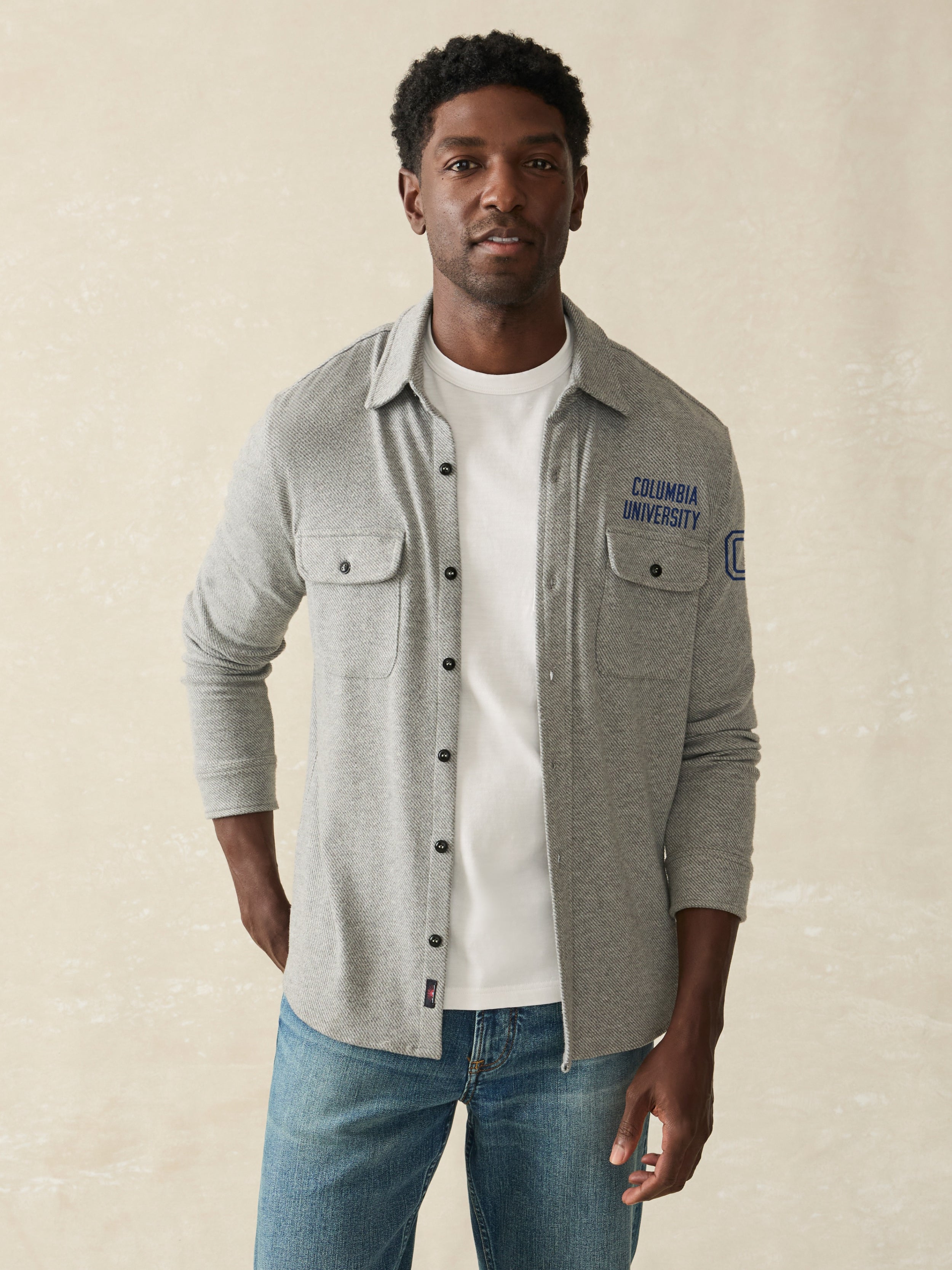 Man wearing a grey long sleeve button down shirt with Columbia University graphic on the left chest and shoulder against a beige background