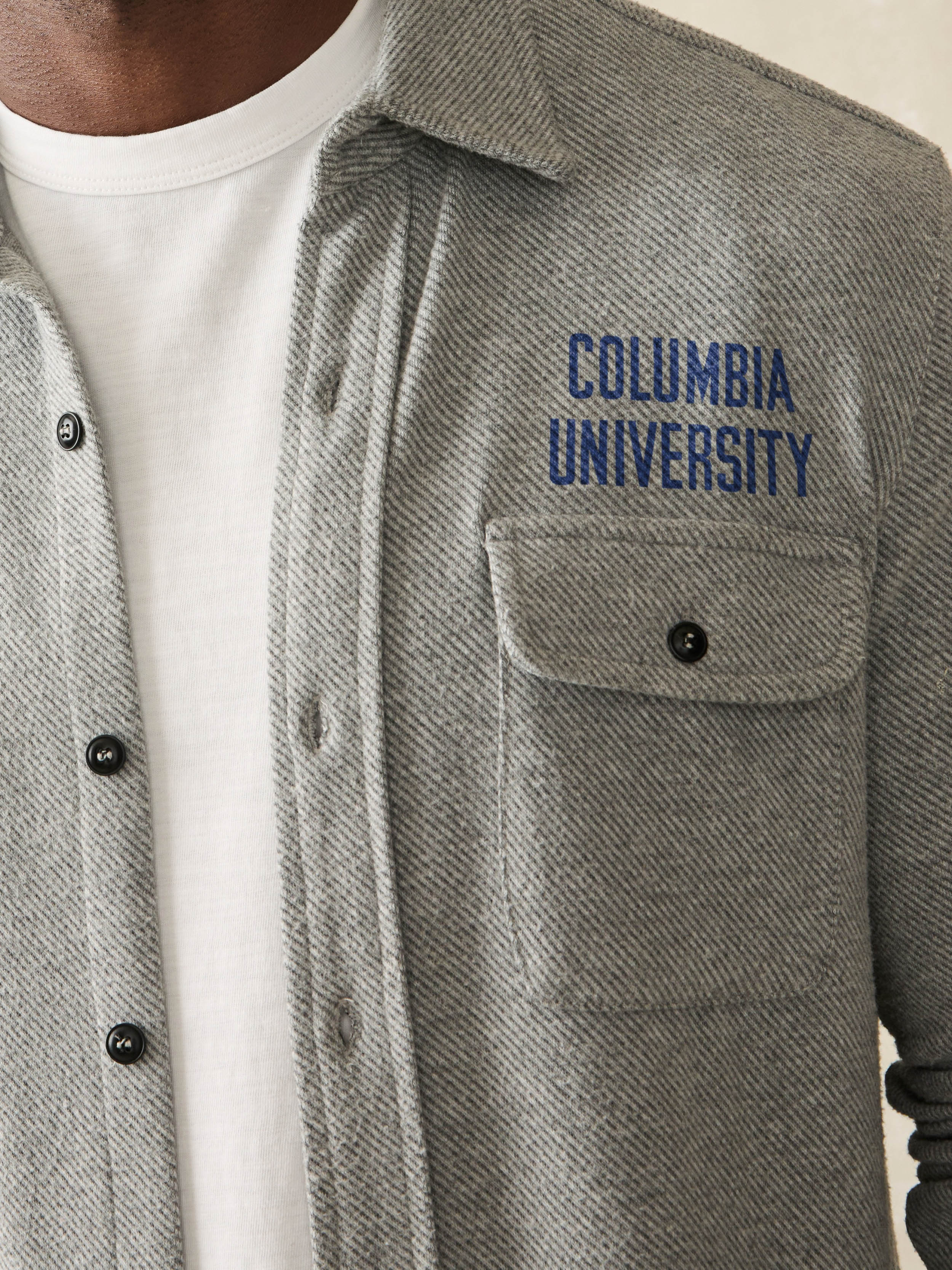 Man wearing a grey long sleeve button down shirt with Columbia University graphic on the left chest and shoulder against a beige background