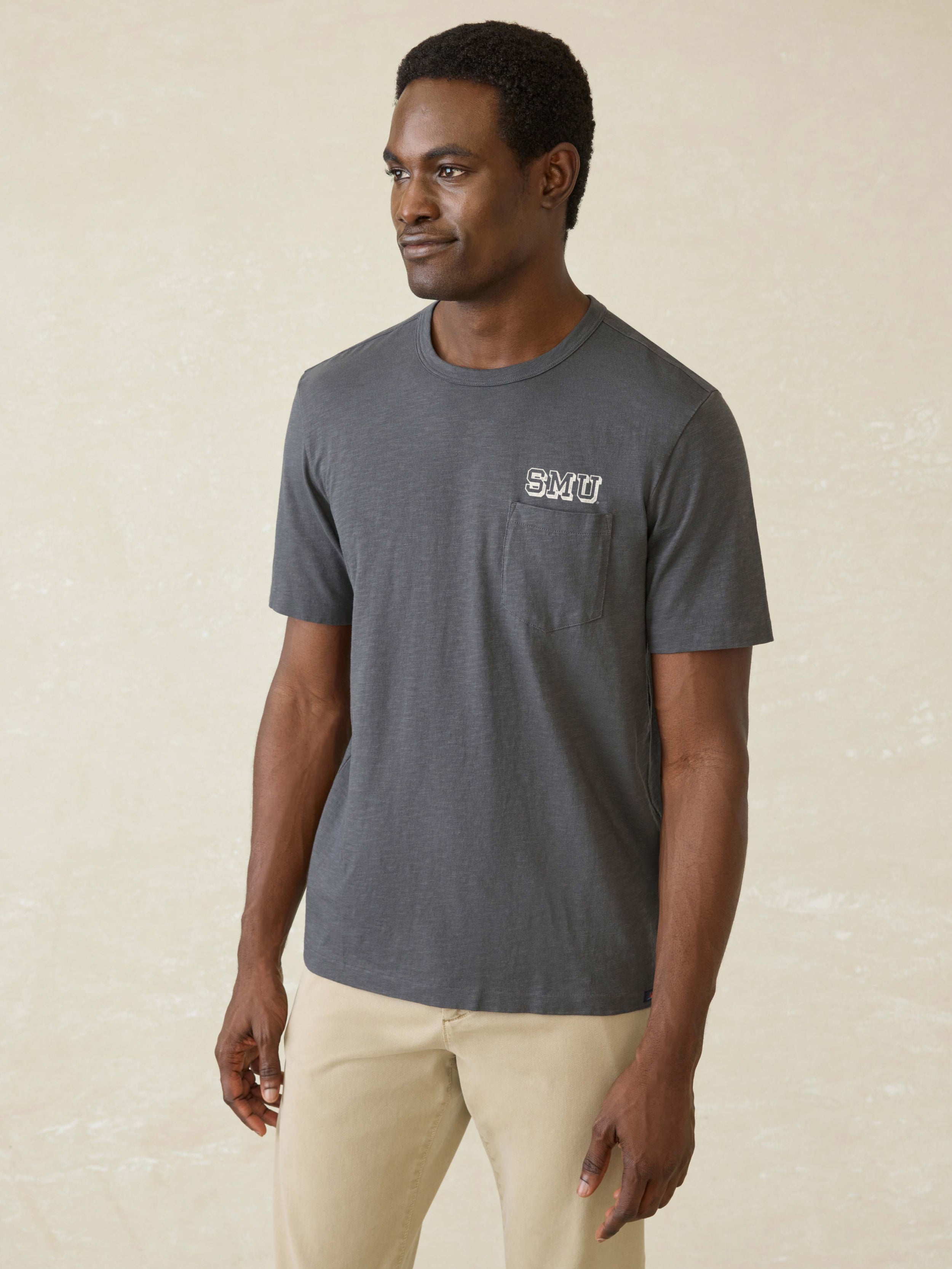 Man wearing a dark grey short sleeve t-shirt with SMU graphic at left chest pocket against a beige background
