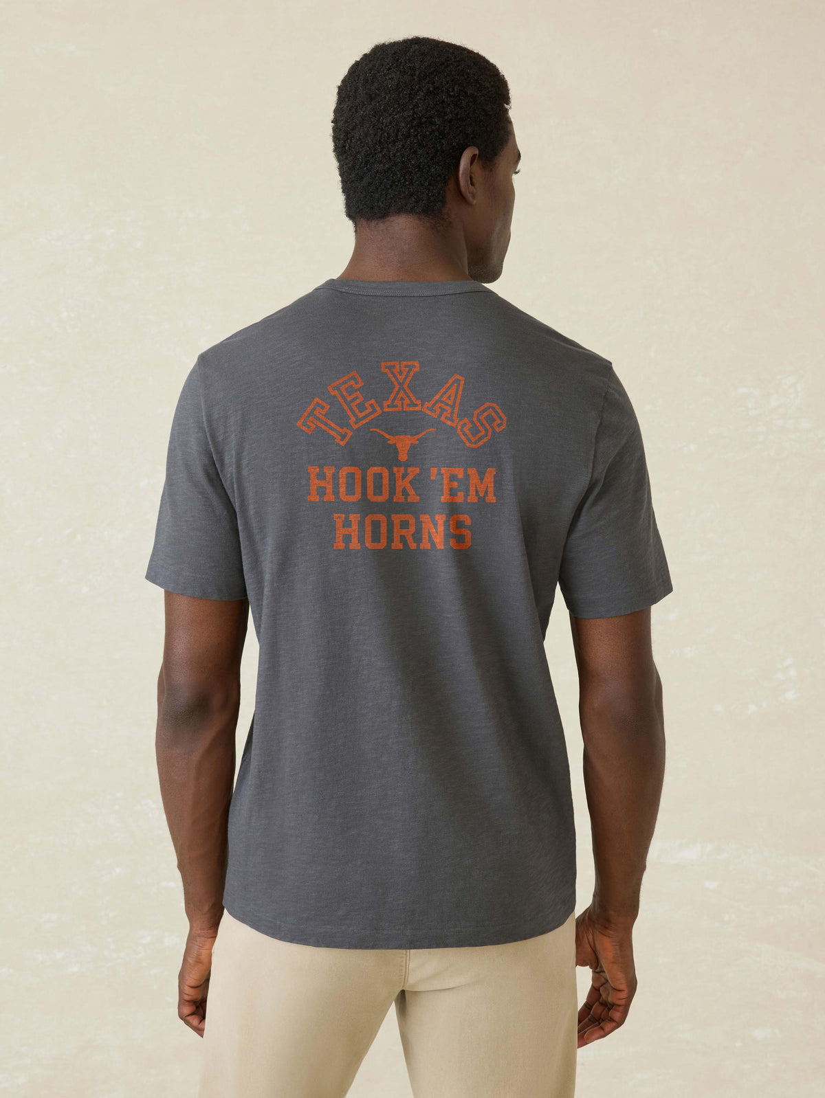 A man viewed from behind wearing a grey University of Texas tee and beige pants, image 2 of 2