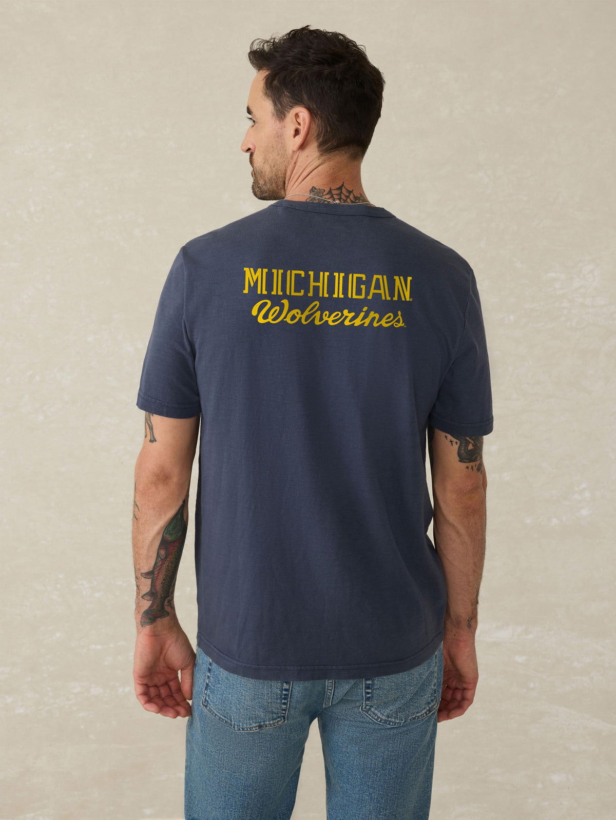 A man viewed from behind wearing a blue University of Michigan tee and blue jeans, image 2 of 2