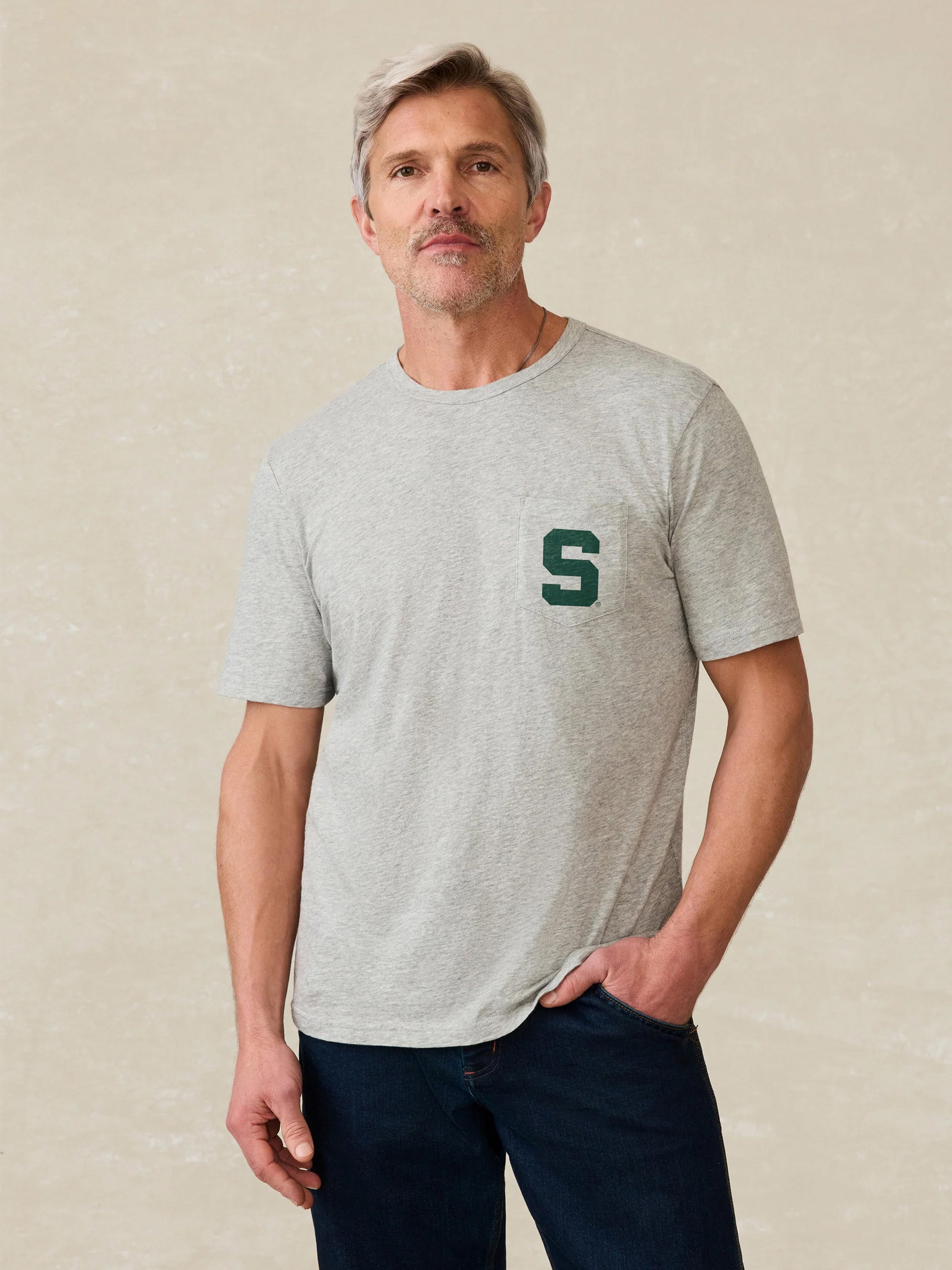 Man wearing a gray t-shirt with a Michigan State graphic logo on the left chest pocket on a beige background