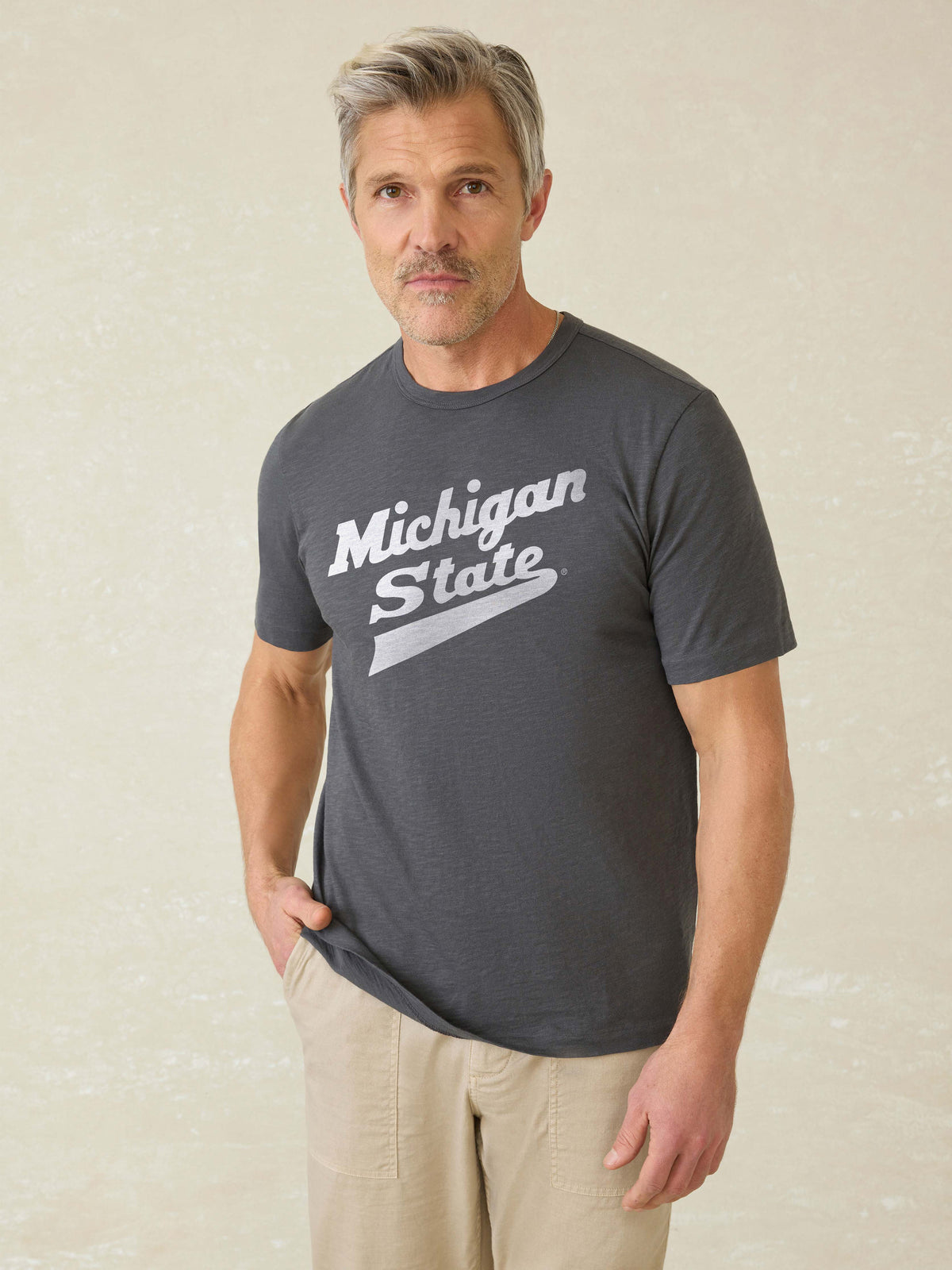 Man wearing a grey t-shirt with Michigan State graphic logo at the center front against a beige background, image 1 of 1