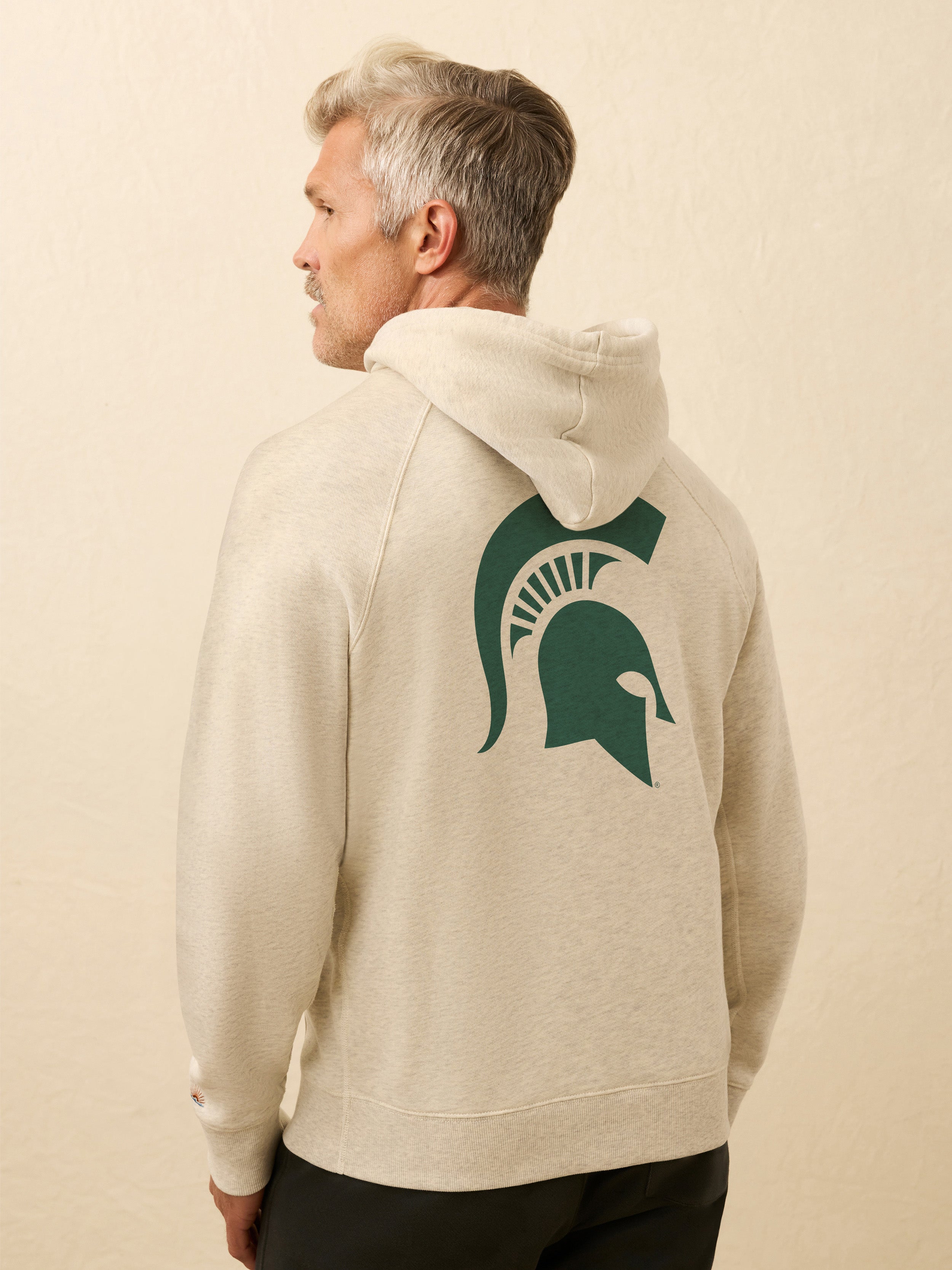 Man wearing a beige hoodie with a green Michigan State graphic on the back against a beige background